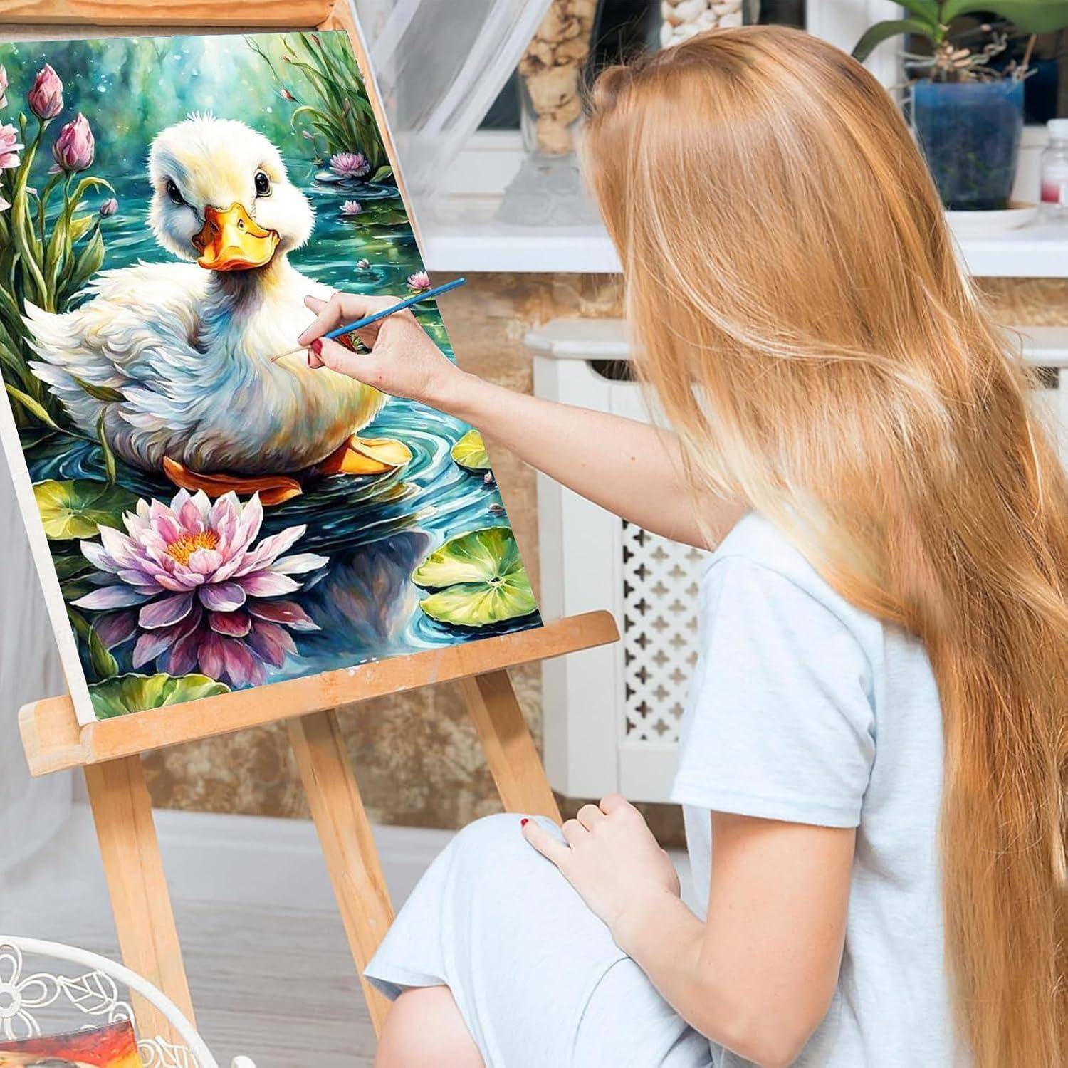 Frau malt ein Entchen auf eine Leinwand an einer Staffelei, mit Seerosen in einem hellen Innenraum-Atelier.