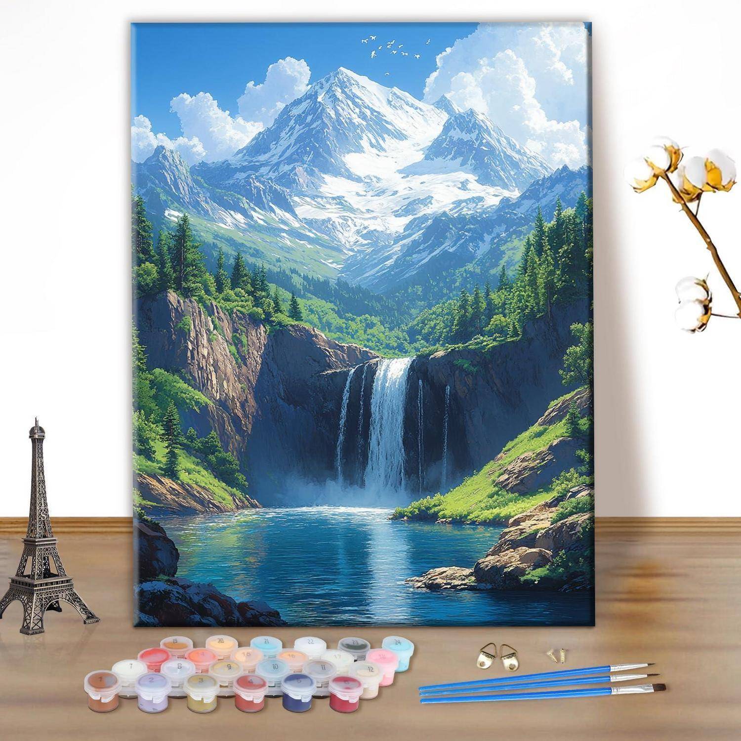 Gerahmtes Gemälde einer Berglandschaft mit verschneitem Gipfel, Wasserfall, Wald und See, ausgestellt auf einem Tisch mit Farben und Pinseln in der Nähe.