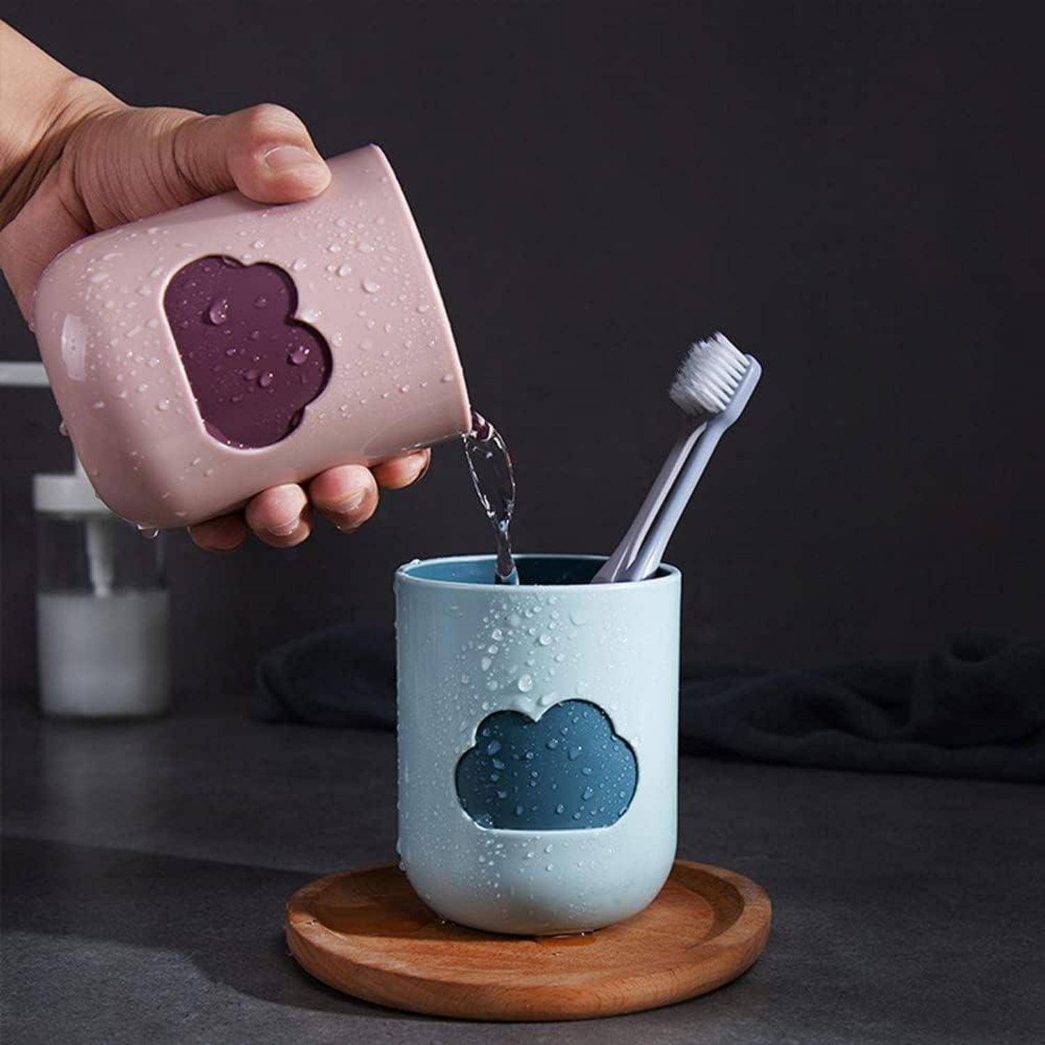 Eine Hand gießt Wasser aus einem rosa wolkenförmigen Behälter in einen passenden blauen Zahnpflegebecher auf einem Holztablett.