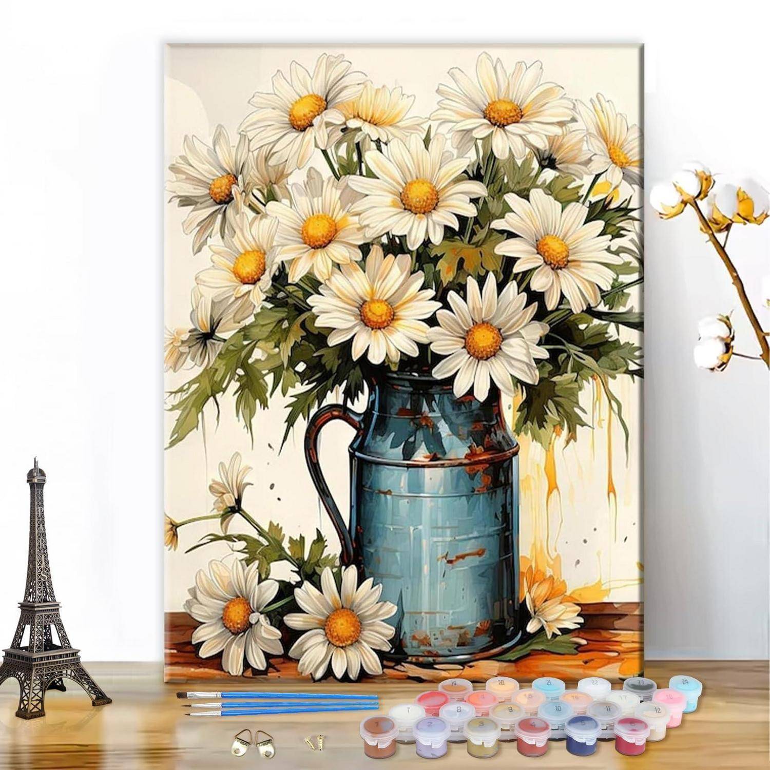 Ein Strauß weißer Gänseblümchen in einem blauen Metallkrug, mit Malutensilien und einer kleinen Eiffelturm-Figur auf einer Tischplatte.