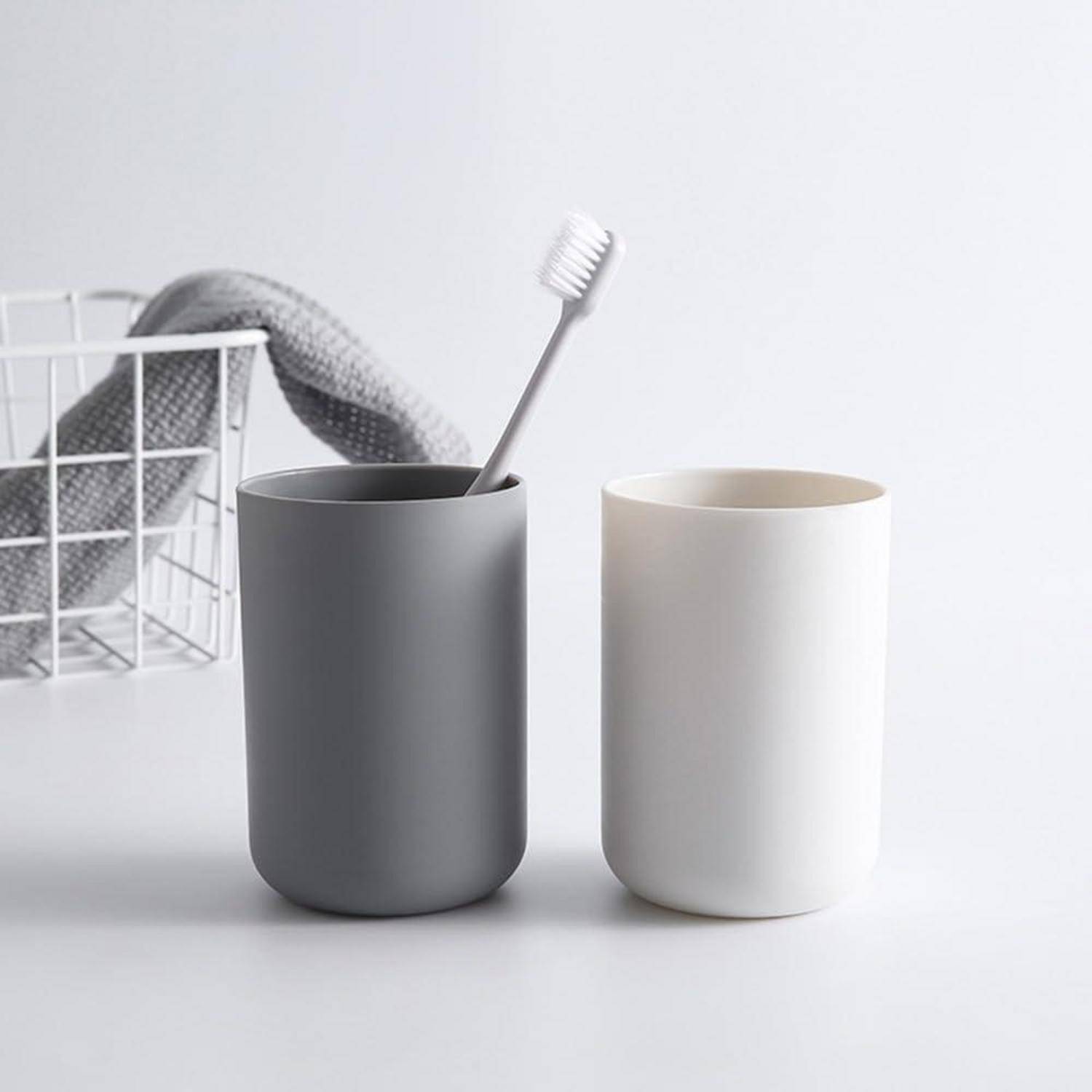 Zwei minimalistische Badezimmergläser, eines grau mit einer Zahnbürste und eines weiß, neben einem Drahtkorb mit einem Handtuch.