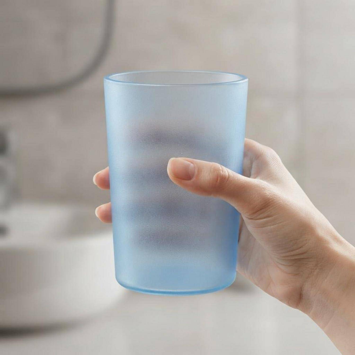 Eine Hand, die einen durchscheinenden, blaugetönten, mattiert Plastikbecher vor einem unscharfen Innenraum-Hintergrund hält.