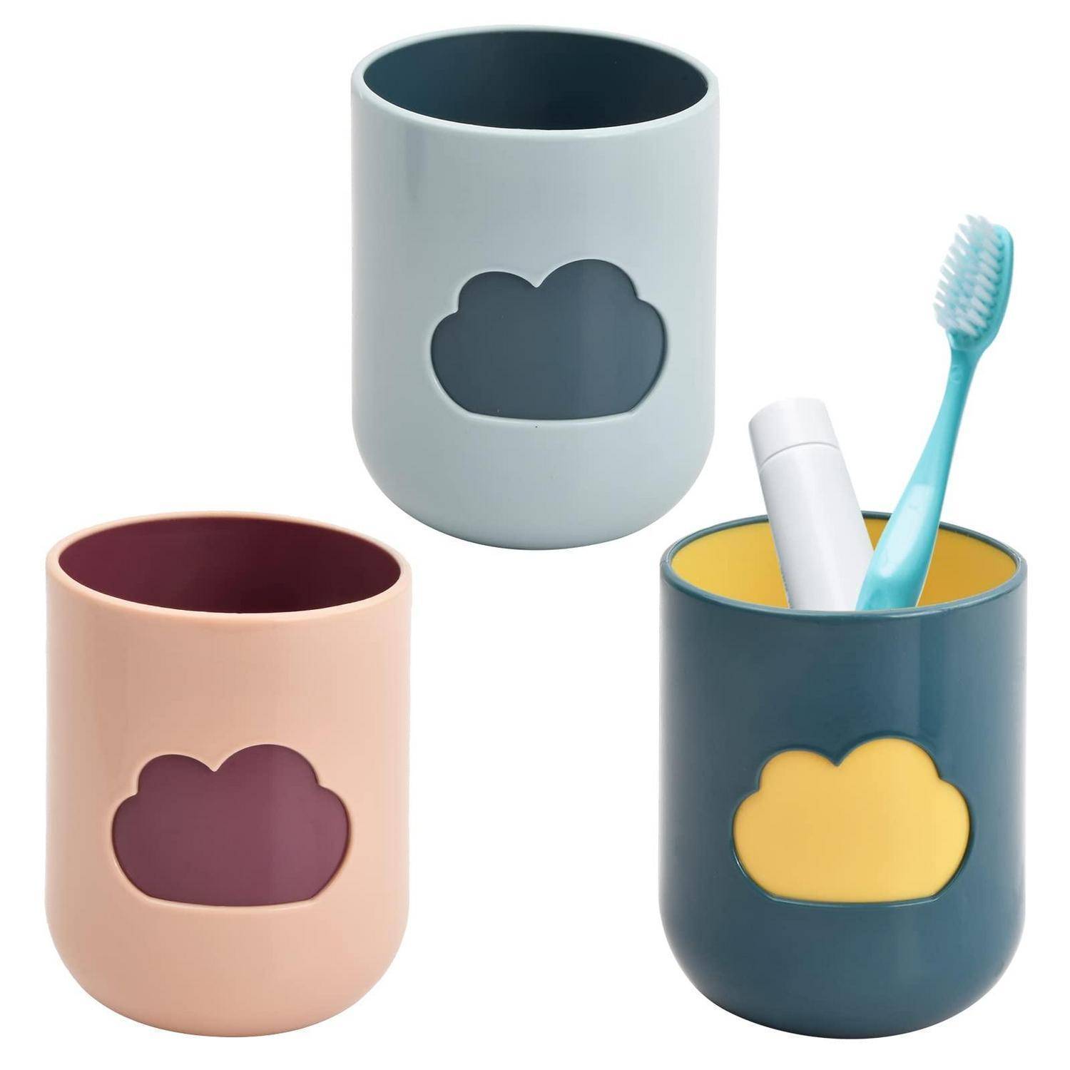 Drei Badezimmertassen im Pastelldesign mit Wolkenmotiv: eine grau, eine rosa und eine dunkelblaugrün, wobei eine Zahnbürste und Zahnpasta hält.