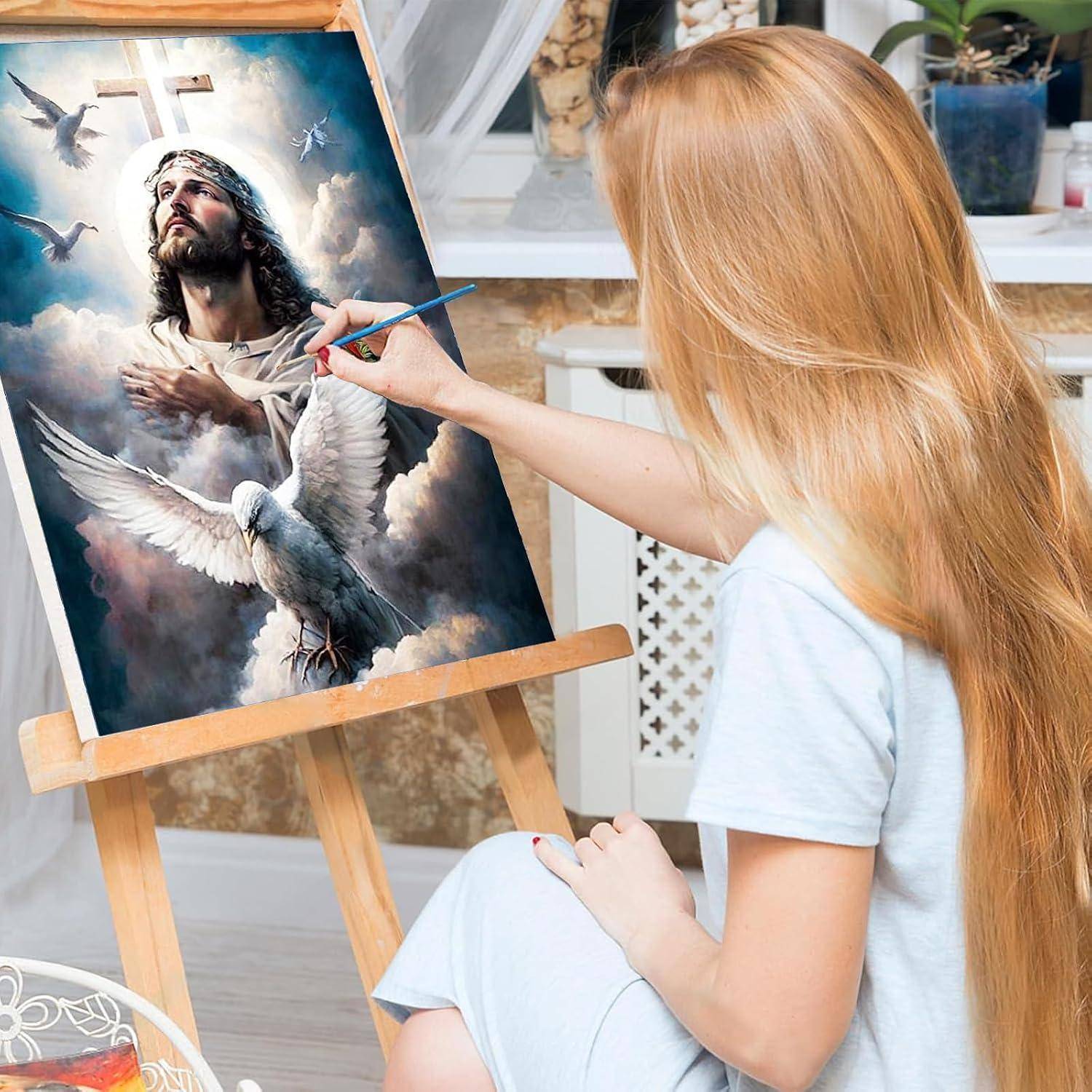 Frau malt eine religiöse Szene auf einer Staffelei, die Jesus mit einem Kreuz und weißen Tauben in einem Atelier darstellt.
