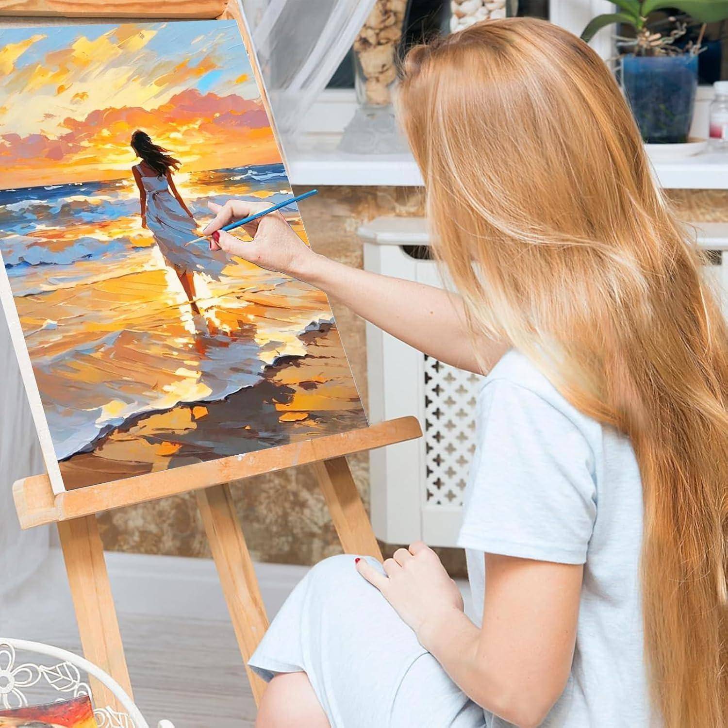 Junge Frau, die auf einer Staffelei in einem Innenraum-Atelier ein Sonnenuntergangs-Seenlandschaftsbild malt und sich dabei auf eine Figur konzentriert, die am Wasser steht.