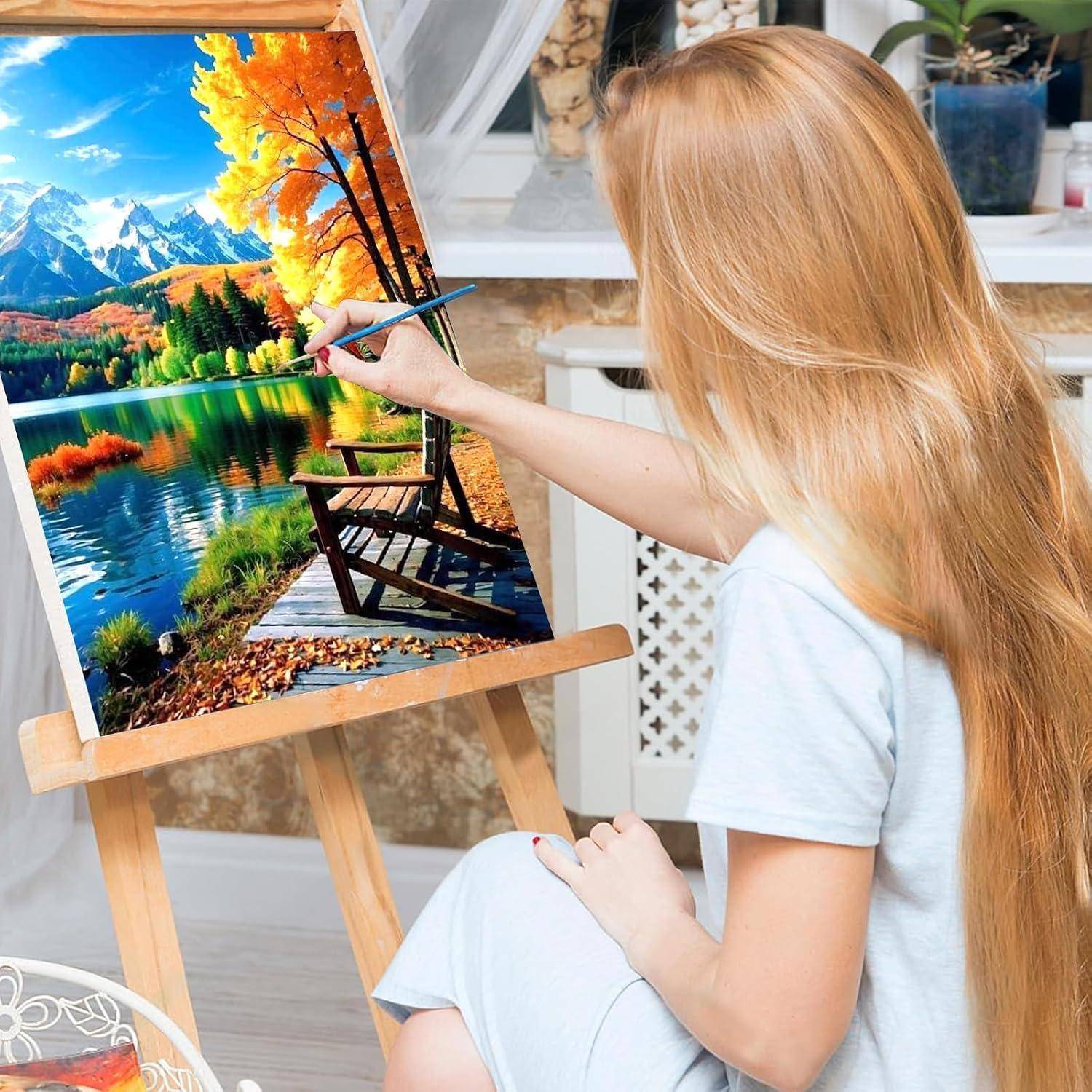 Eine Frau malt eine lebendige Landschaft auf einer Staffelei und fügt Details zu einer See- und Berglandschaft in einer Studioeinstellung hinzu.