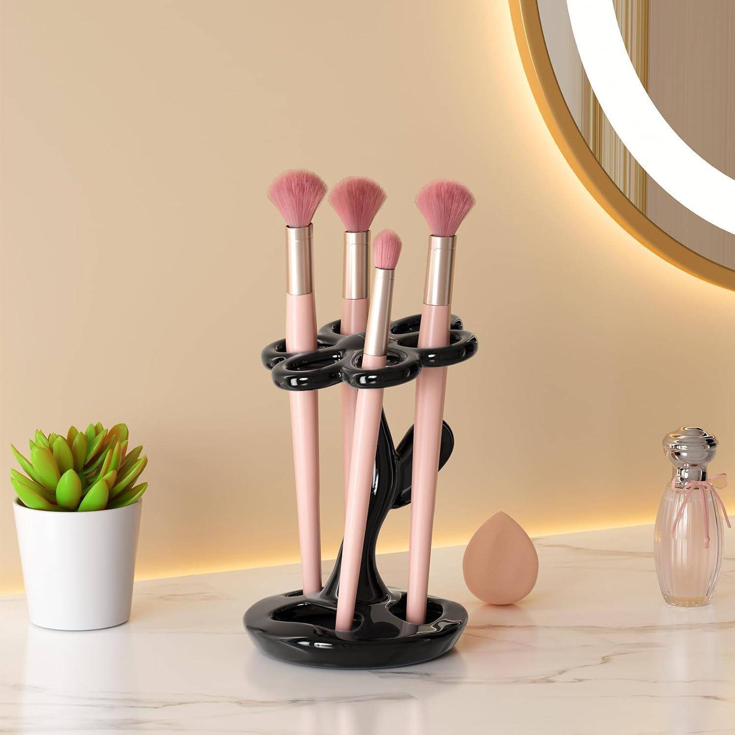 Vier rosa Make-up-Pinsel in einem schwarzen Ständer auf einer Frisierkommode, mit einem Schwamm, einer kleinen Pflanze und einer Parfümflasche in der Nähe.