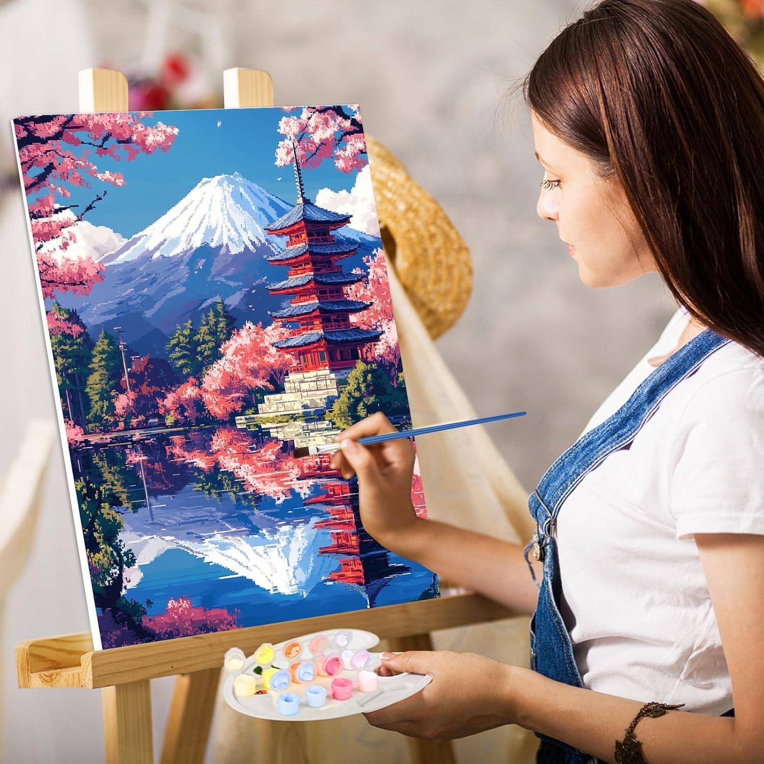 Eine Frau malt eine farbenfrohe Landschaft auf einer Staffelei, die den Berg Fuji, Kirschblüten und eine Pagode mit Wasserreflektion darstellt.