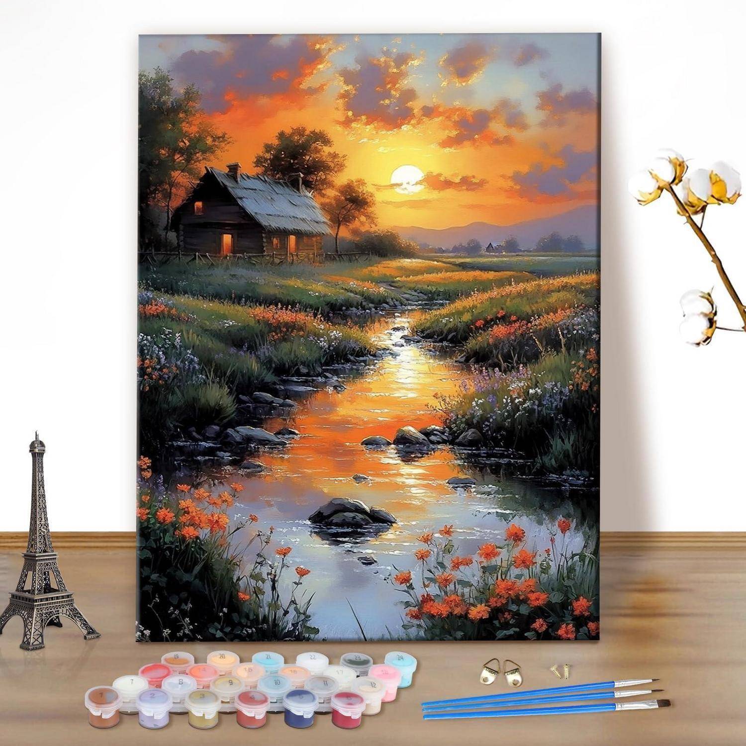 Kunstmaterialien arrangiert vor einer gemalten Sonnenunterganglandschaft einer Hütte an einem Bach, mit einer Eiffelturm-Figur und Blumen im Vordergrund.