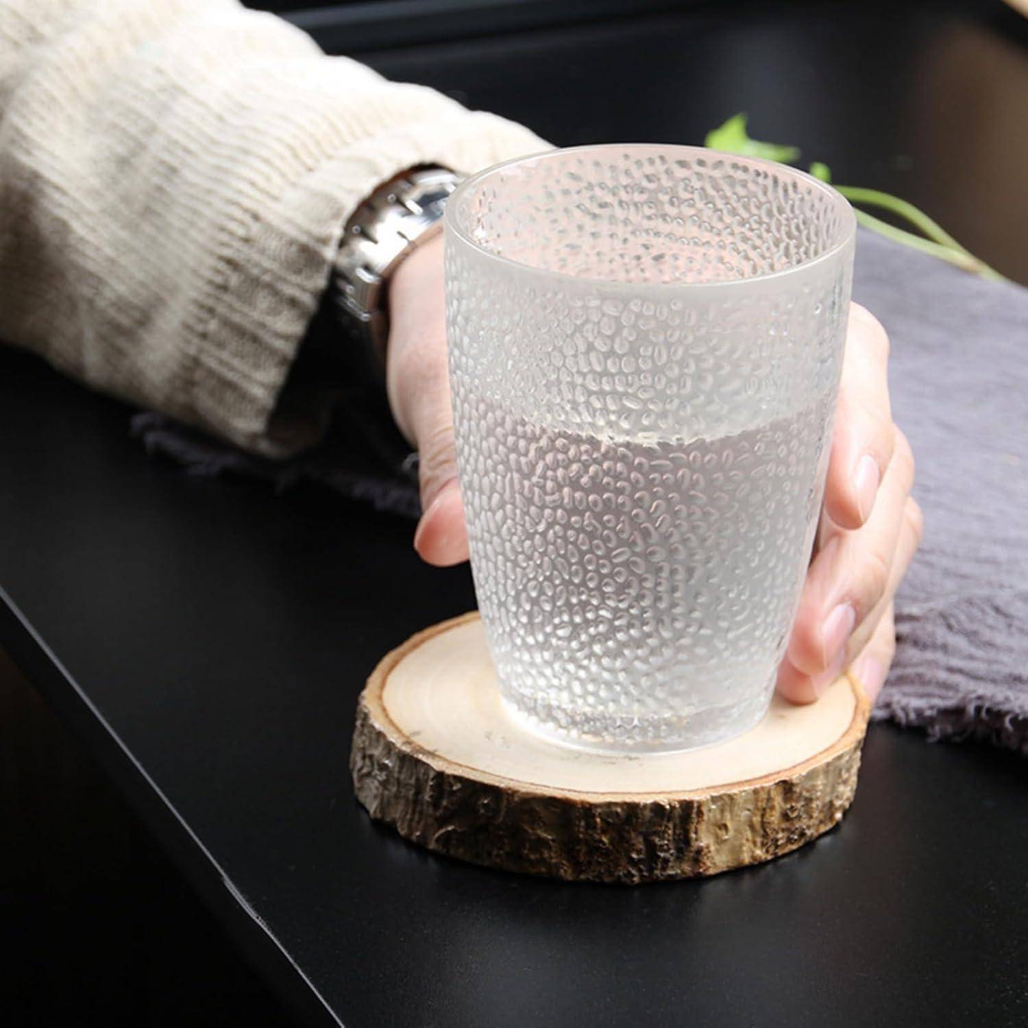 Eine Hand hält ein Glas Wasser mit Reifbildung auf einem Holzuntersetzer auf einem schwarzen Tisch.