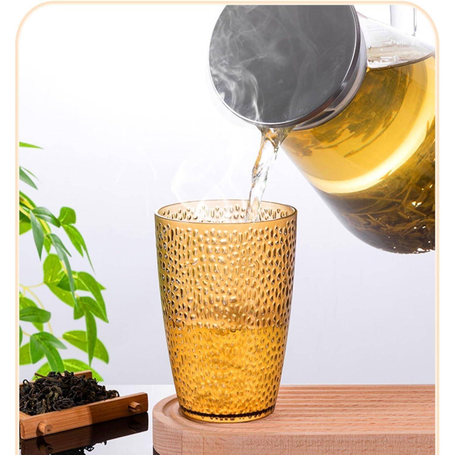 Klarer Tee wird aus einer Glaskanne in ein bernsteinfarbenes strukturiertes Glas gegossen, neben grünen Blättern und losem Tee auf einem Holztablett.