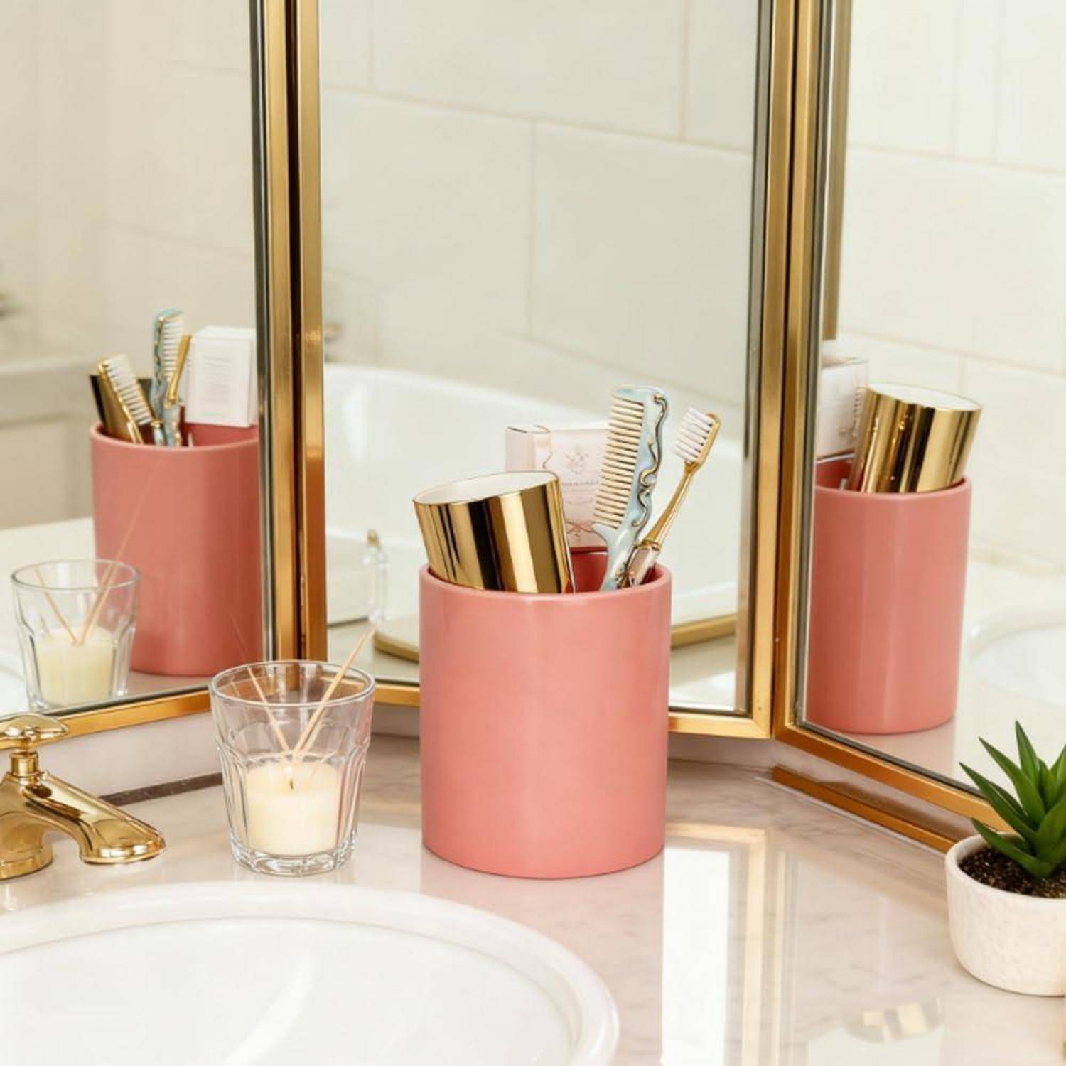 Badezimmerschrank mit rosa Organizer, die Zahnbürsten und Toilettenartikel neben einem Waschbecken halten, eine Kerze, einen goldenen Wasserhahn und eine kleine Pflanze.