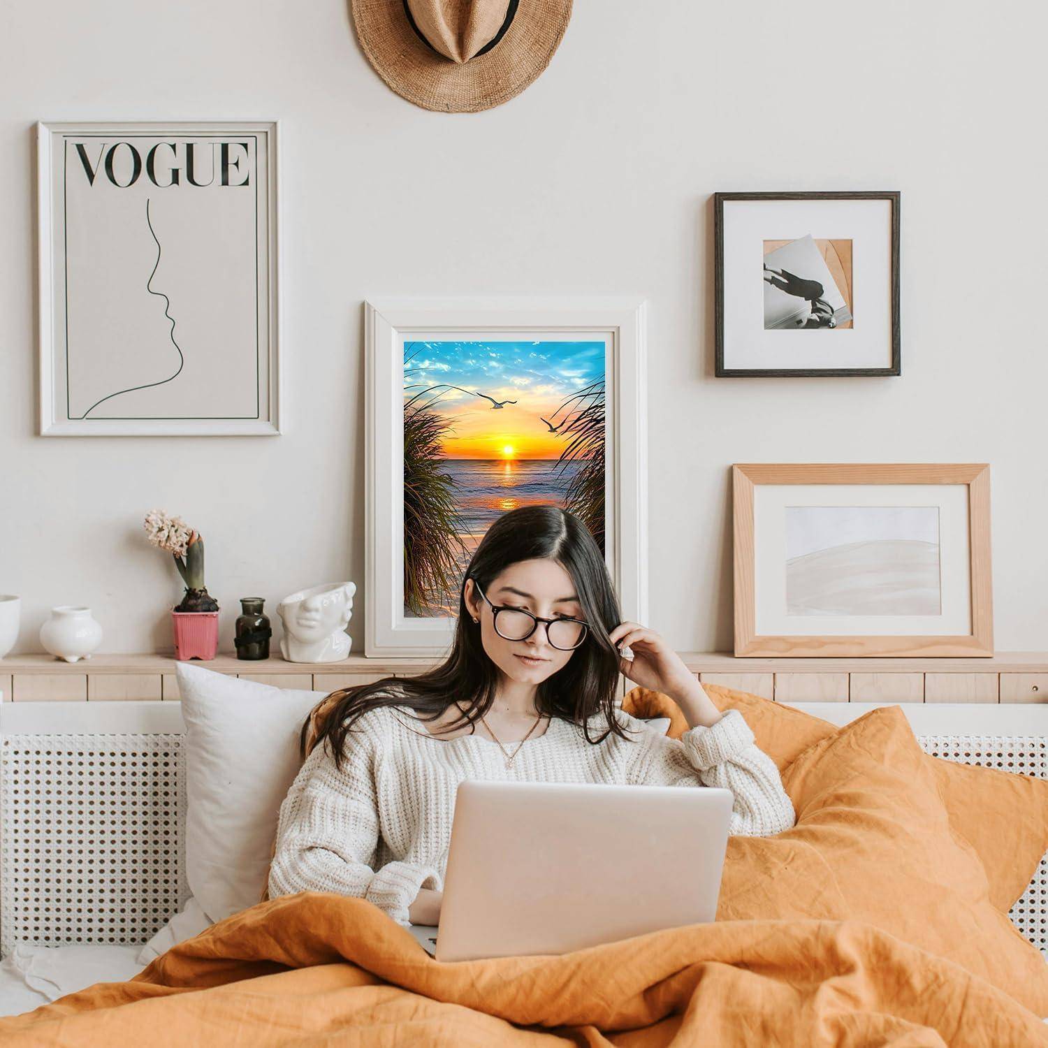 Junge Frau im Bett, die einen Laptop benutzt, mit gerahmter Wandkunst hinter ihr und einem zentrierten Sonnenuntergangsstrandbild über dem Kopfende.