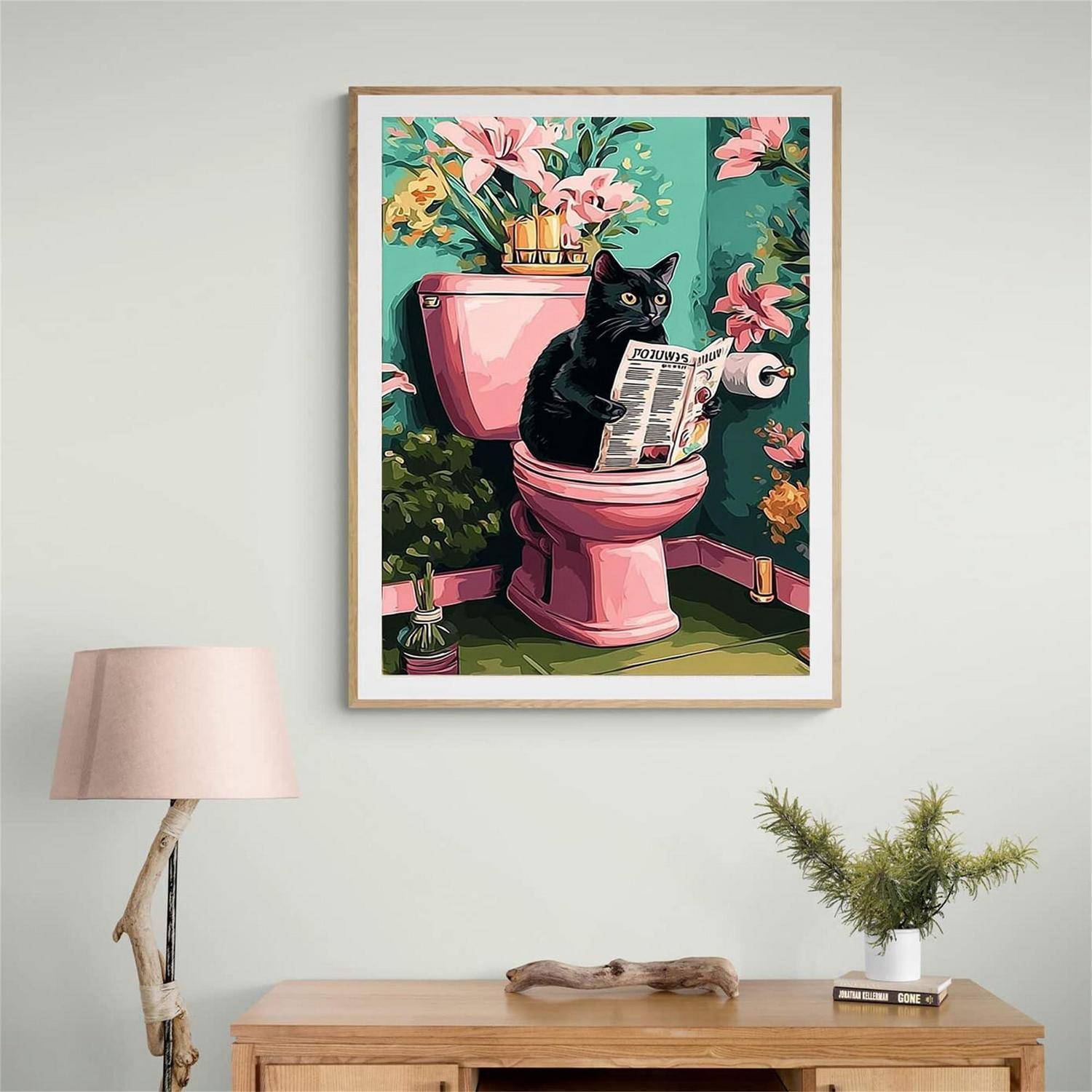 Gerahmtes Kunstwerk einer schwarzen Katze, die eine Zeitung liest, während sie auf einer rosa Toilette sitzt, an einer Wand über einem Holzkonsoltisch angebracht.