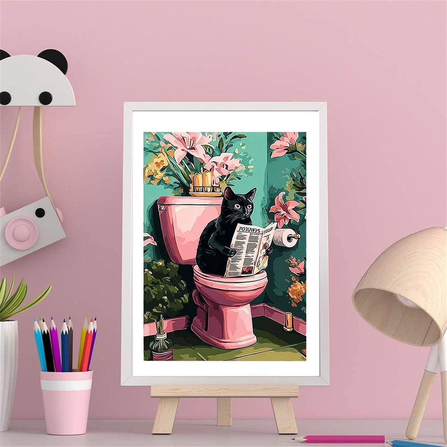 Gerahmte Illustration einer schwarzen Katze, die eine Zeitung liest, während sie auf einer rosa Toilette sitzt, ausgestellt auf einer Staffelei in einem rosa Zimmer.