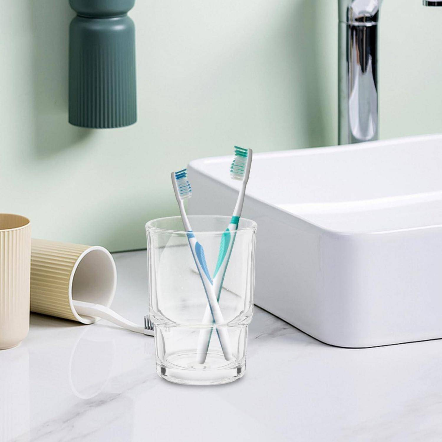 Zwei Zahnbürsten in einem durchsichtigen Glas neben einem weißen Badezimmerspülbecken und einer Tasse, mit einer mintgrünen Wand und einem Chromhahn im Hintergrund.