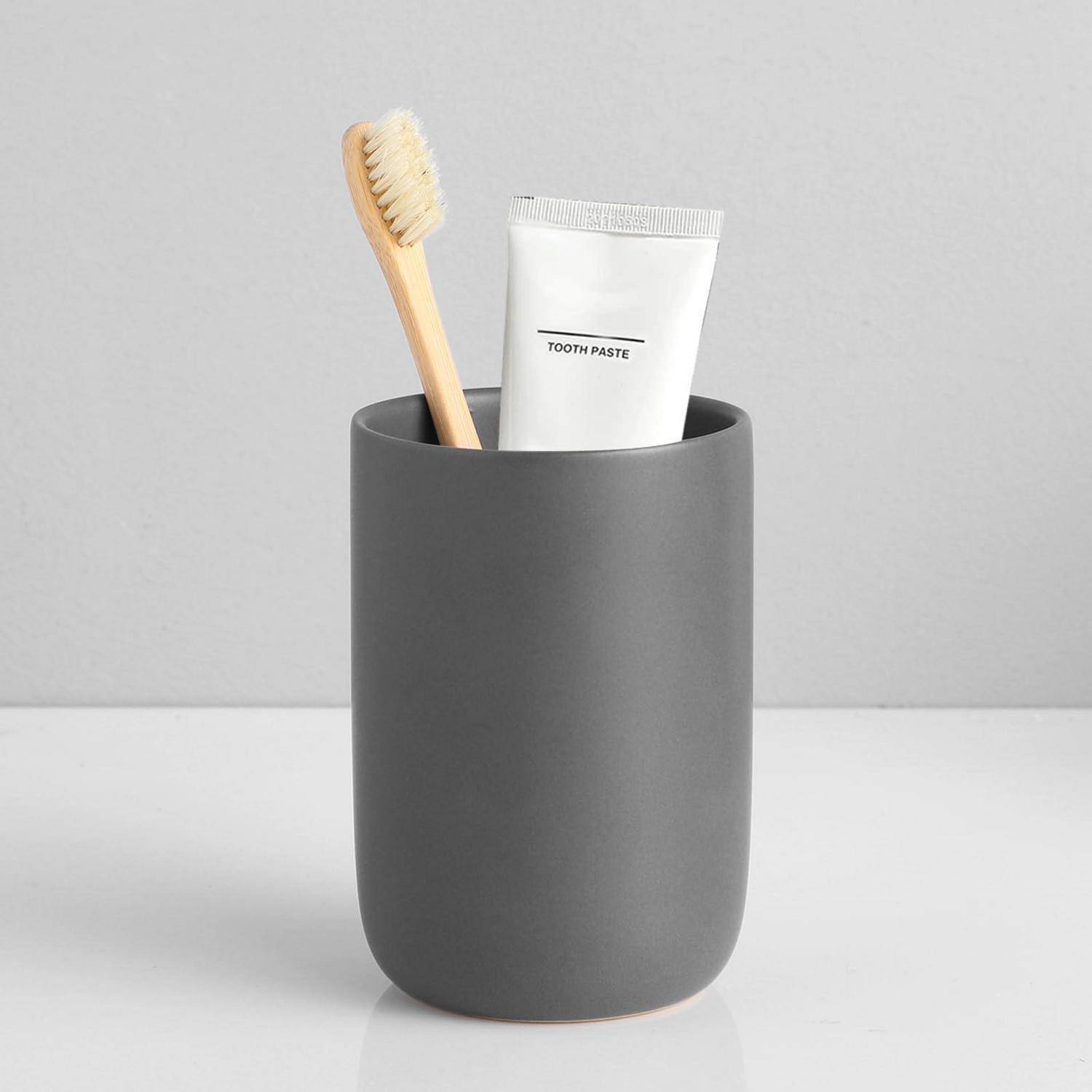 Ein grauer Badezimmertasse hält eine hölzerne Zahnbürste und eine weiße Zahnpastatube mit der Aufschrift „TOOTH PASTE