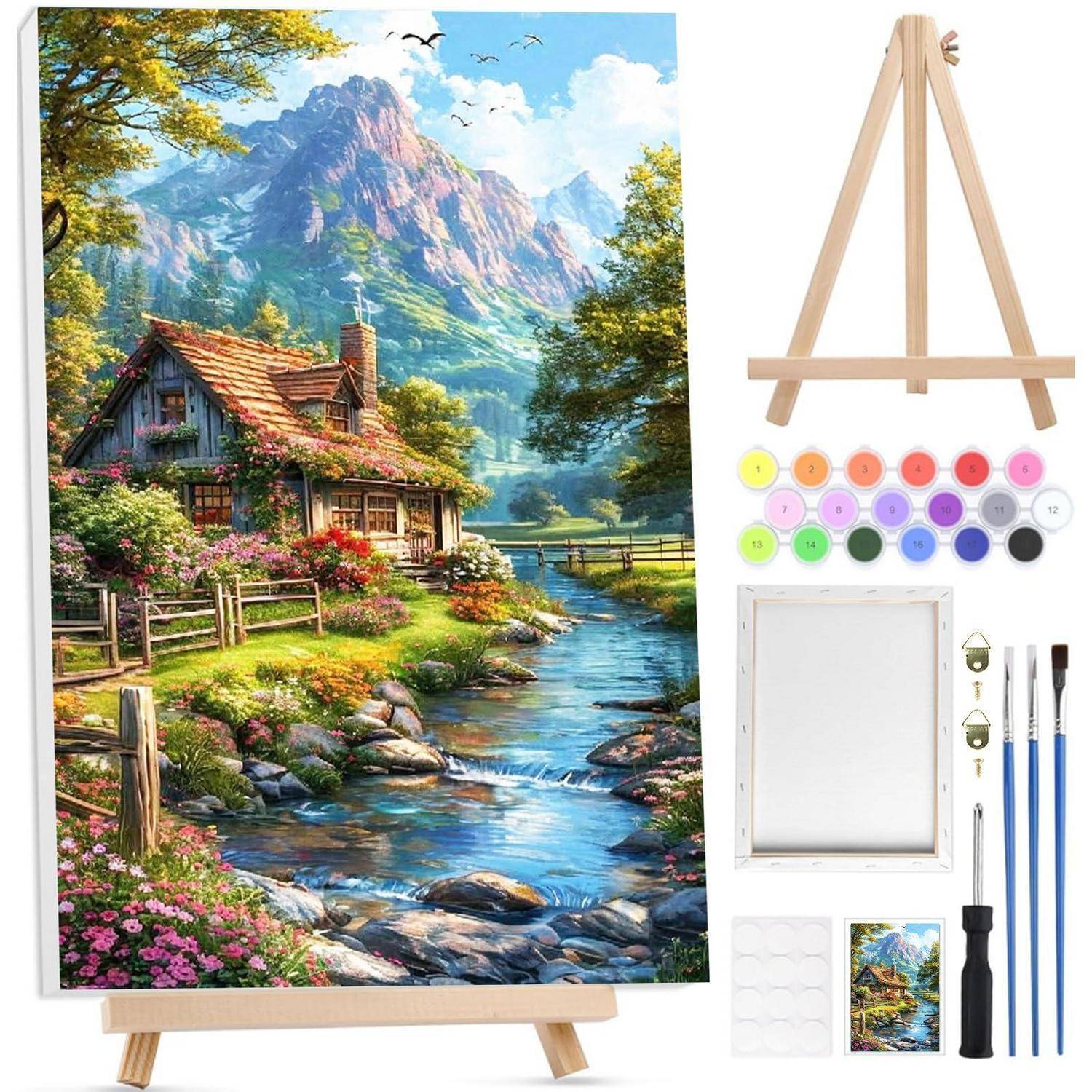 Malerset mit leeiner Leinwand auf einer Staffelei, Farben und Pinseln neben einem Referenzbild einer Berghütte an einem Bach