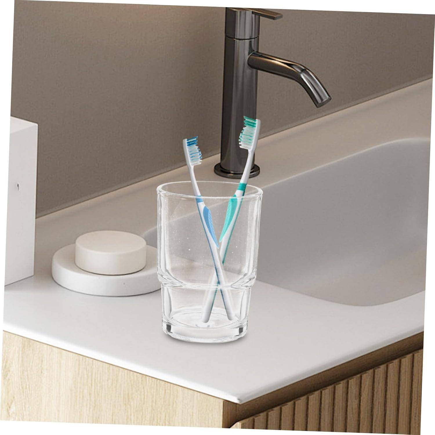 Zwei Zahnbürsten in einem klaren Glas neben einem Badezimmerspülbecken und einem Wasserhahn.