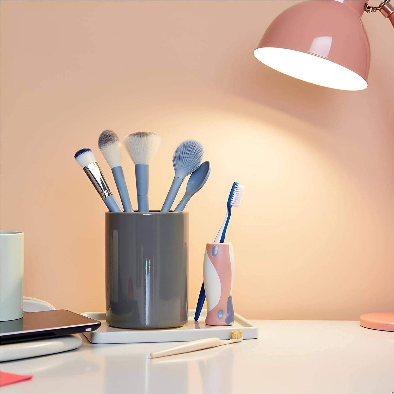 Schreibtisch mit einer grauen Tasse mit Makeup-Pinseln, einer Zahnbürste in einem rosa Halter, einer Lampe und einem geschlossenen Laptop auf einer ordentlichen weißen Oberfläche.