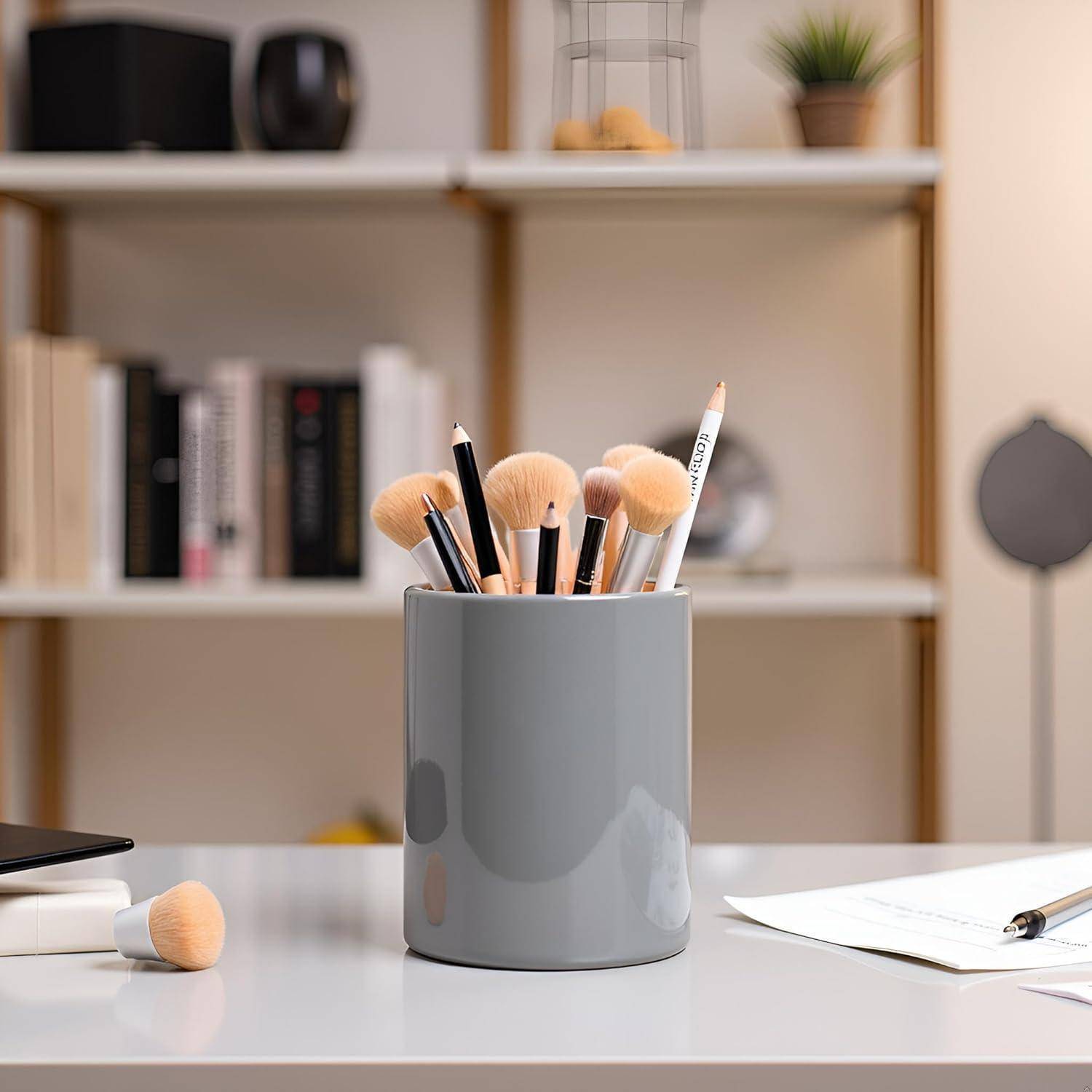Grauer Becher mit Makeup-Pinseln und Stiften auf einem Schreibtisch, mit einem Pinsel, Papieren und Regalen im Hintergrund.