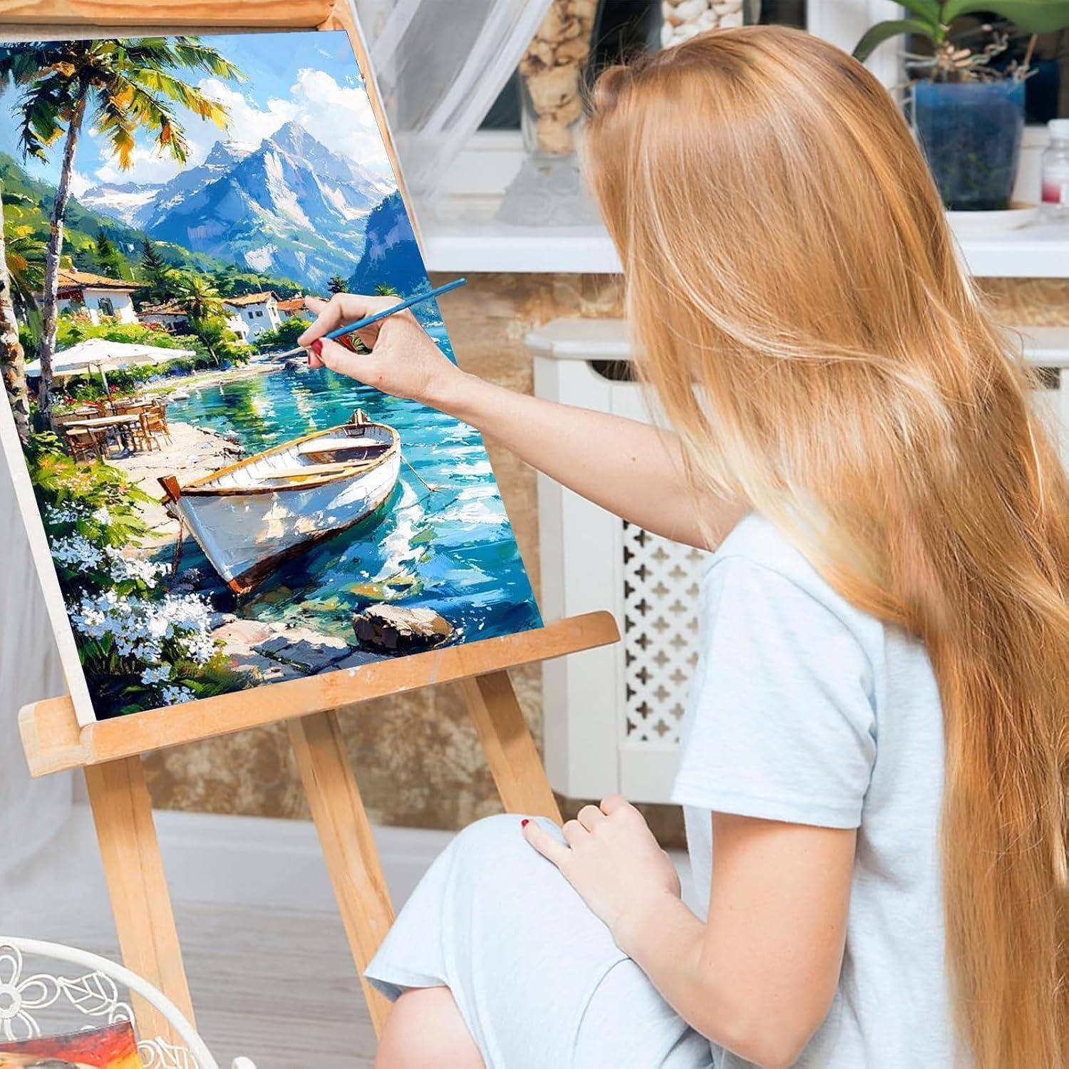 Blonde Frau, die eine lebendige Seeufer-Berglandschaft mit einem Boot auf einer Leinwand an einer Staffelei im Innenbereich malt.