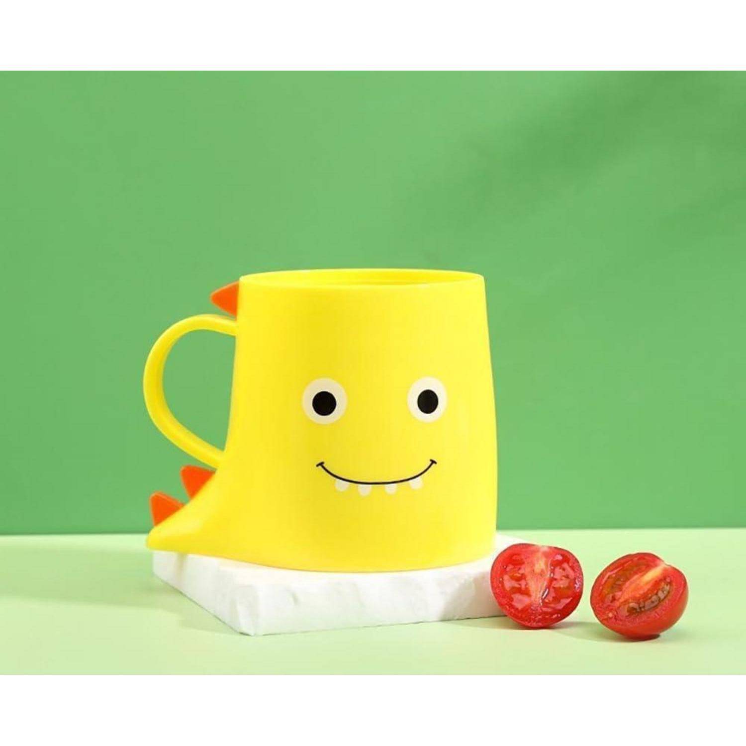 Gelbe dinosaurierförmige Tasse mit lachendem Gesicht, die auf einer weißen Basis neben zwei halbierten Kirschtomaten auf grünem Hintergrund sitzt.