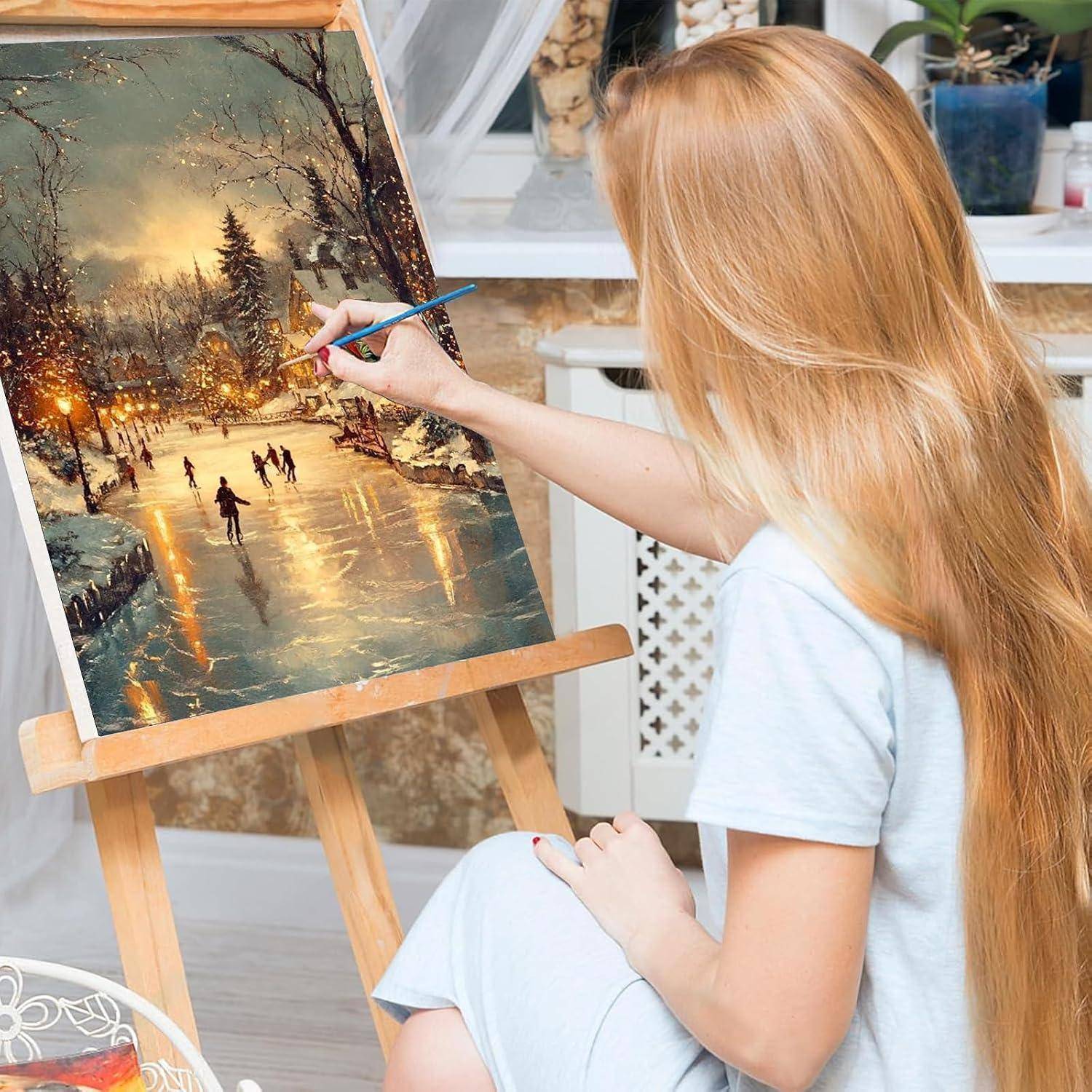 Eine Frau malt eine Winterlandschaft auf einer Staffelei in einem hellen Raum und fügt Details zu einer verschneiten Parkszene mit Schlittschuhläufern und warmen Lichtern hinzu.