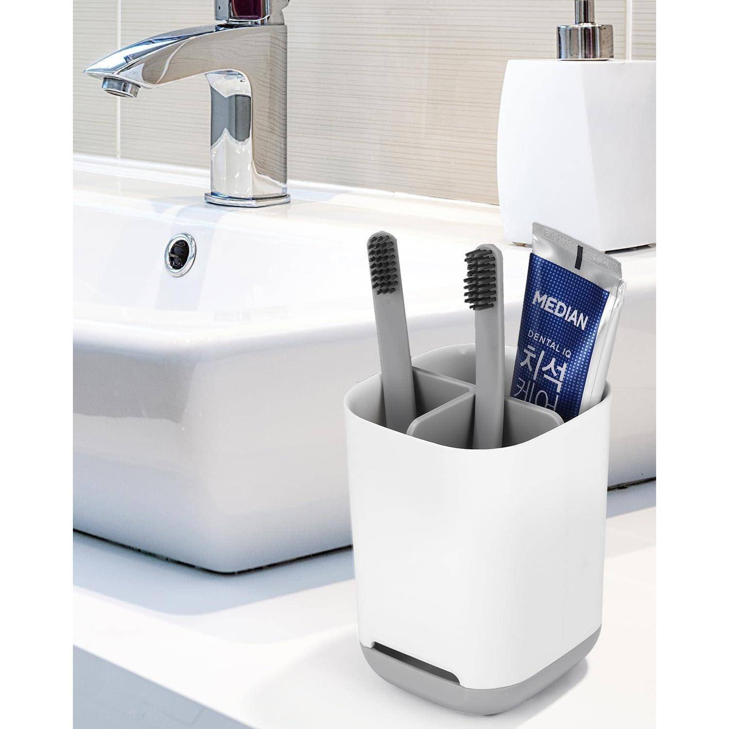 Zahnbürstenhalter mit zwei Zahnbürsten und einer Zahnpastatube neben einem weißen Badezimmerbecken und einem Wasserhahn.