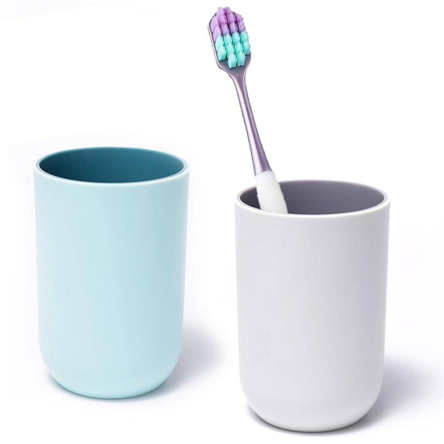 Zwei minimalistische Badezimmertassen auf weißem Hintergrund, eine hellblau und eine weiß, mit je einer lila Zahnbürste darin.