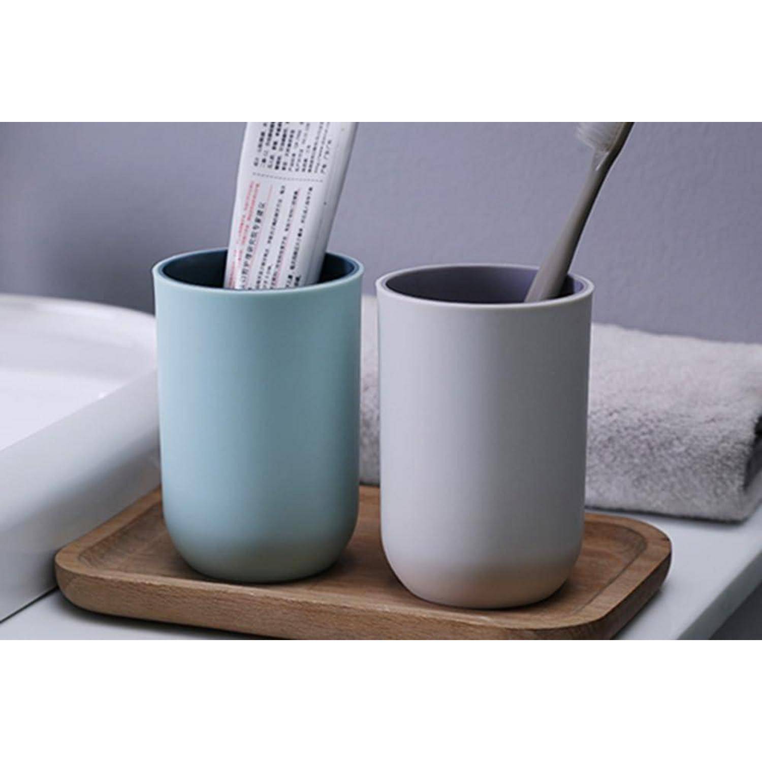 Zwei minimalistische Badezimmertassen auf einem Holztablett neben einem Waschbecken, eine mit einer Zahnbürste und einer Tube, mit einem gefalteten Handtuch im Hintergrund.