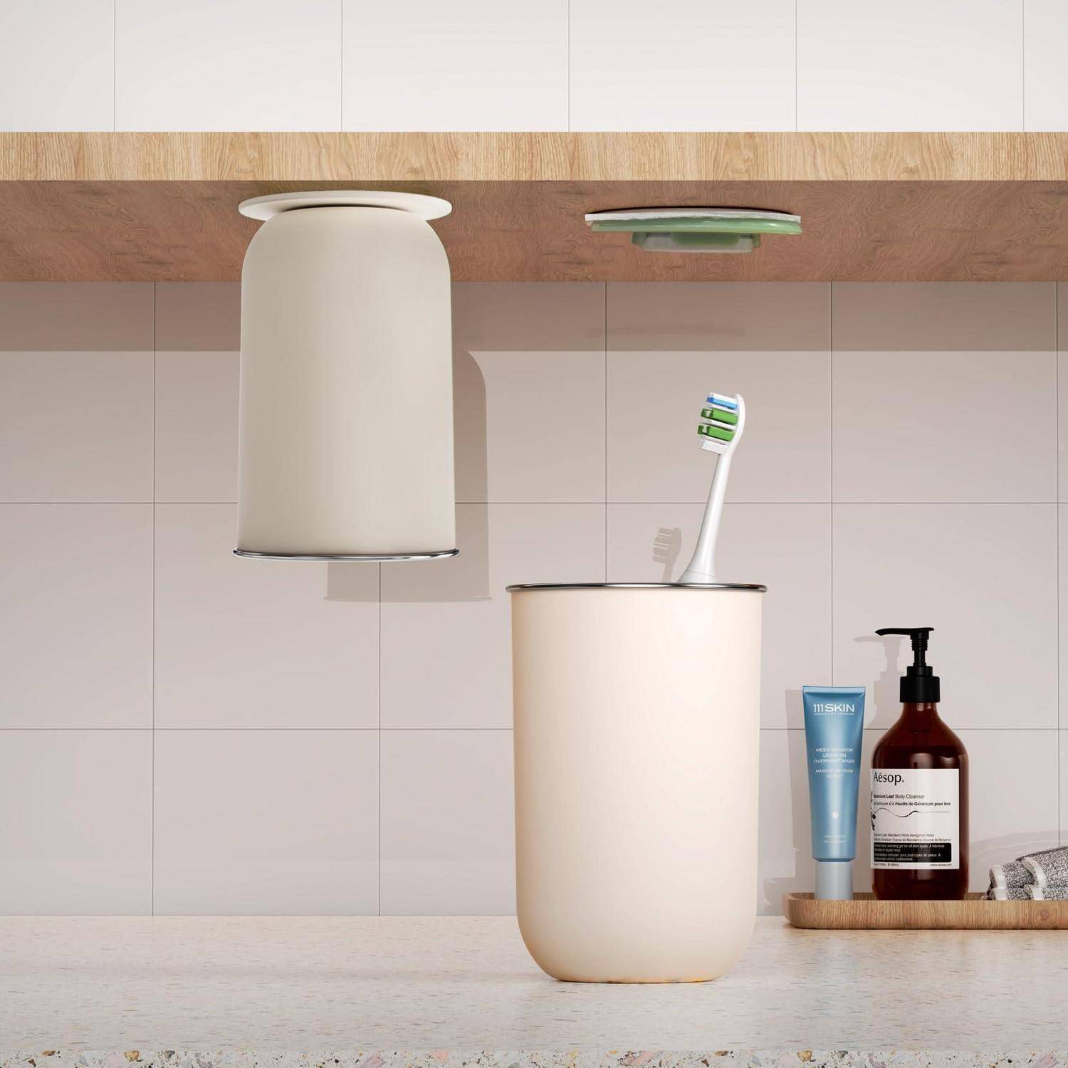 Minimalistischer Badezimmertisch mit einer Zahnbürste in einem cremefarbenen Becher, einem entsprechenden wandmontierten Behälter darüber und Seifenprodukten neben einem Waschbecken.
