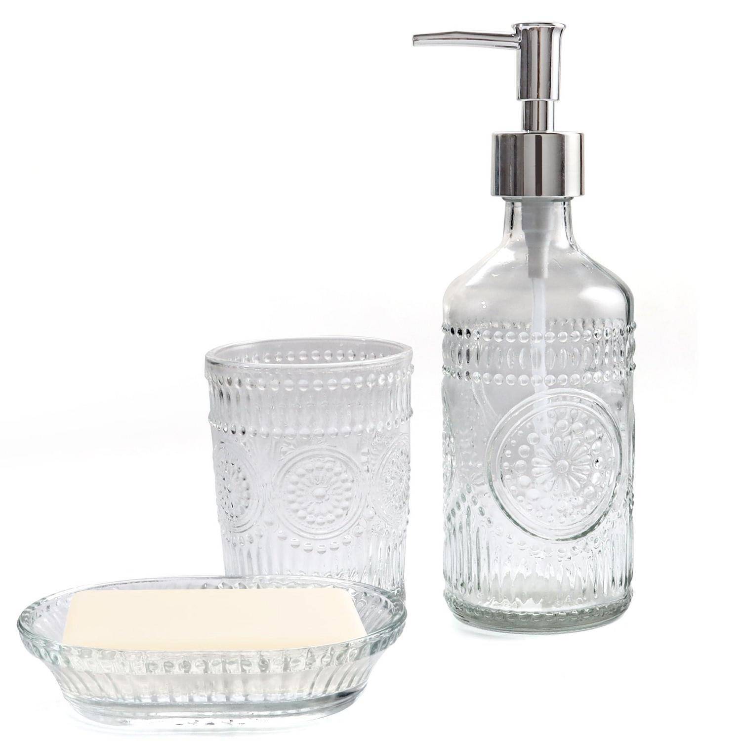 Drei durchsichtige Badezimmeraccessoires aus Glas: ein Seifenspender, ein Zahnputzbecher und eine Seifenschale mit geprägtem Blumenmuster.