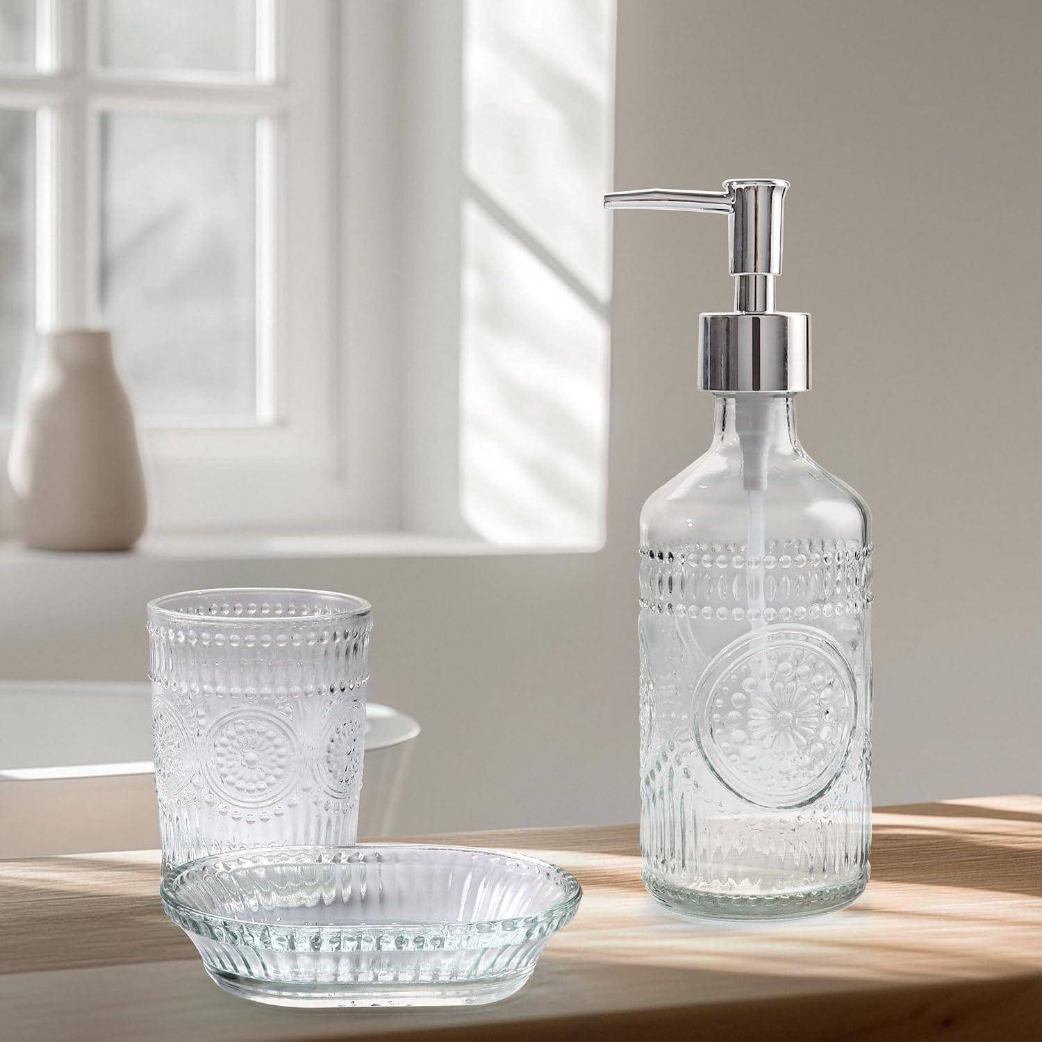 Klare Glasseifenspender mit Pumpe, passender Becher und kleine Schale auf einem sonnigen Holztisch in der Nähe eines Fensters.