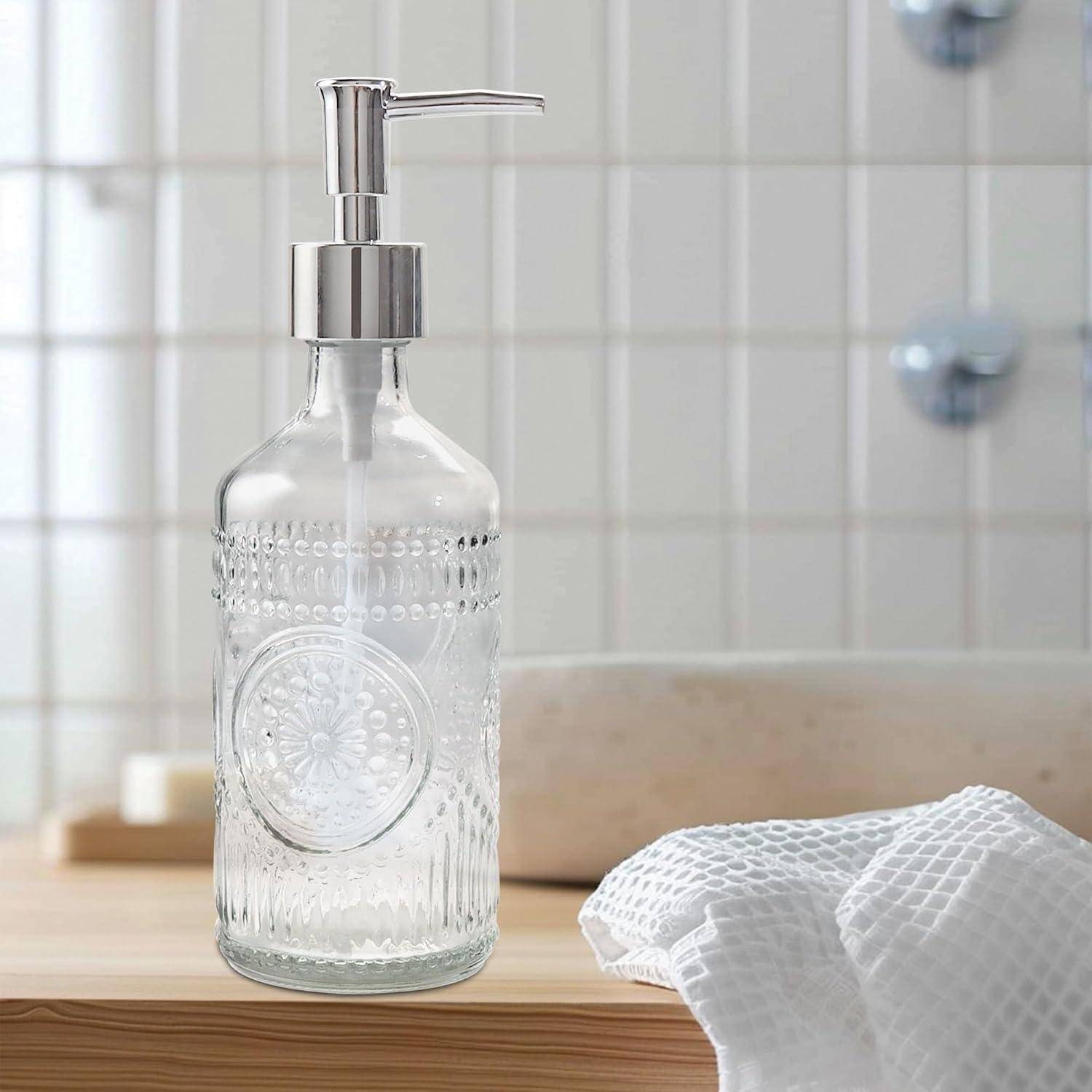 Dekorativer Glasseitenspender mit Metallpumpe auf einer Badezimmerablage neben einem weißen Handtuch und einer Spülenrückwand.