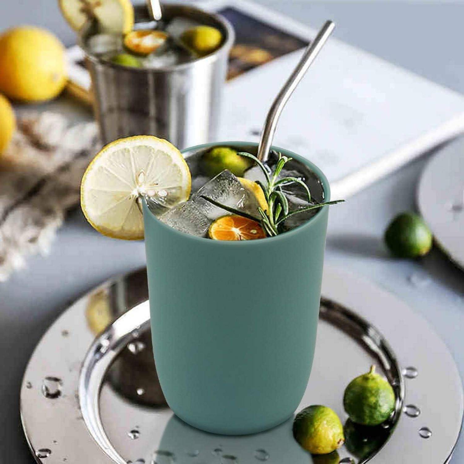 Ein blaugrüner Cocktail in einer mattierten Tasse mit Eis, Zitronenscheibe, Kräutern und kleinen Zitrusgarnituren, serviert auf einem Teller mit einem Strohhalm.