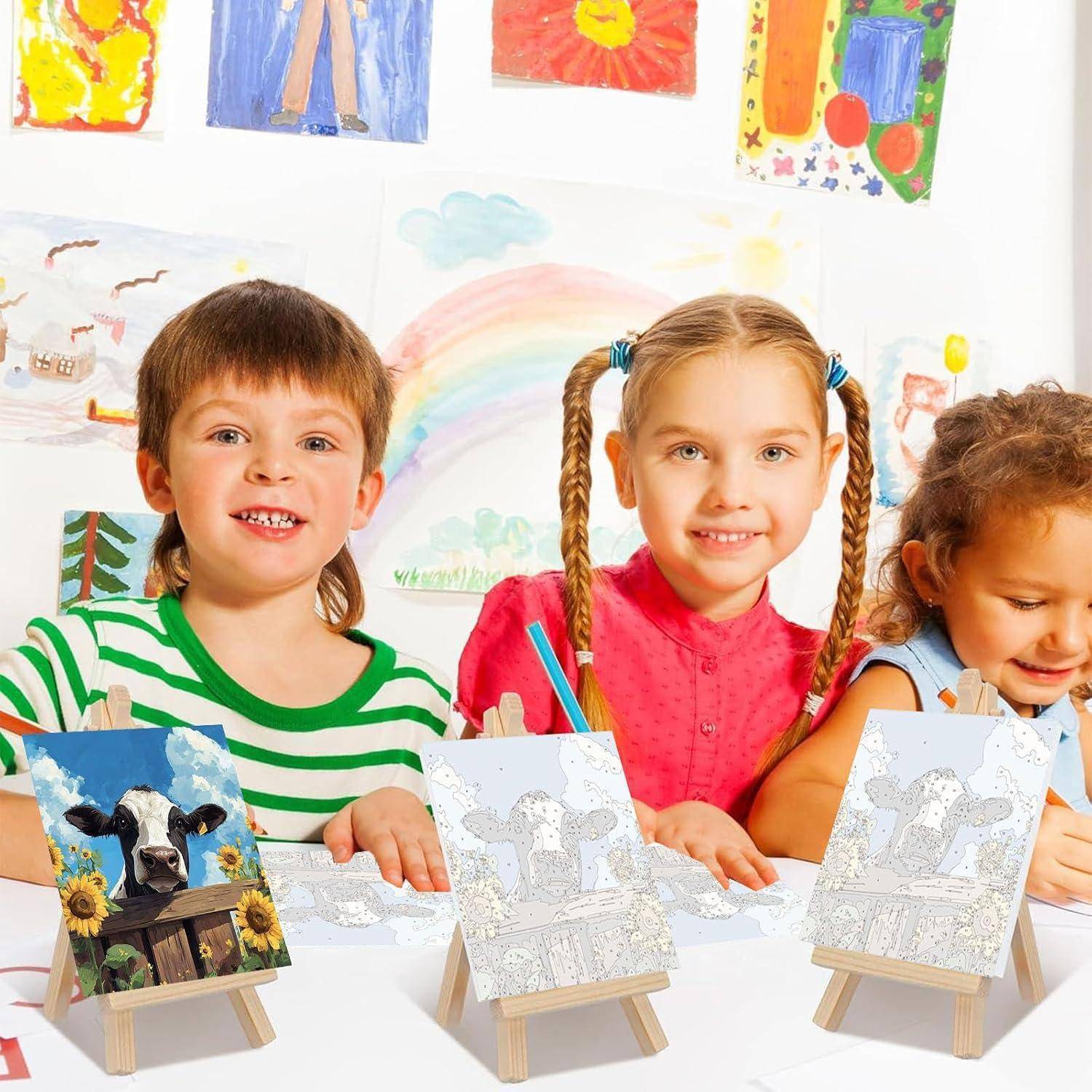 Drei kleine Kinder sitzen an einem Tisch in einem Klassenzimmer und malen kleine Kuhbilder auf Staffeleien, wobei bunte Kinderkunstwerke an der Wand hinter ihnen hängen.