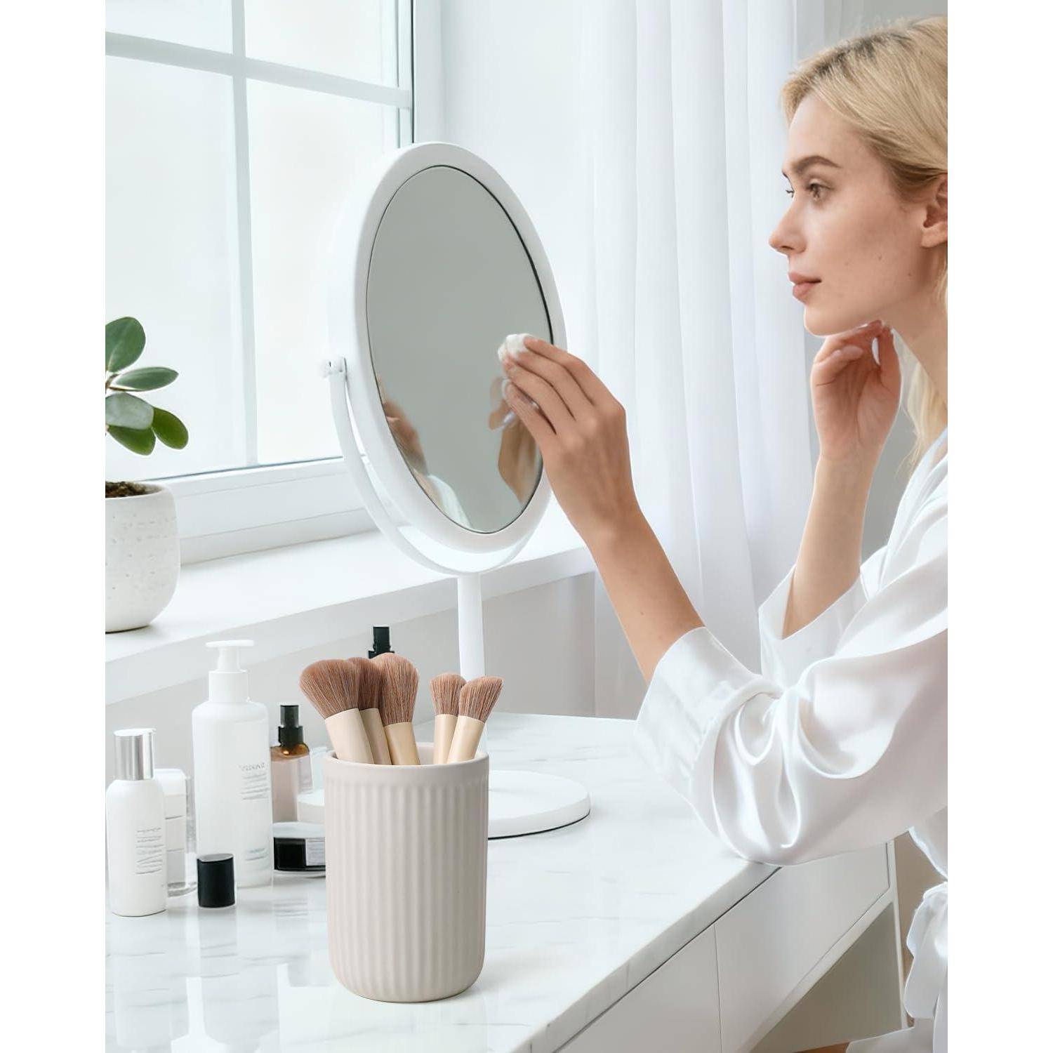 Frau in einem weißen Bademantel trägt Make-up auf, während sie in einen Tischspiegel schaut, neben Kosmetikprodukten und Pinseln bei einem hellen Fenster.