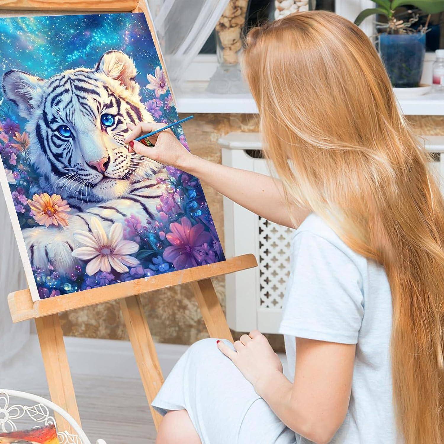 Eine Frau malt einen blauäugigen weißen Tiger auf einer Staffelei und fügt Details zu einem farbenfrohen Blumenkunstwerk in einem Atelier hinzu.