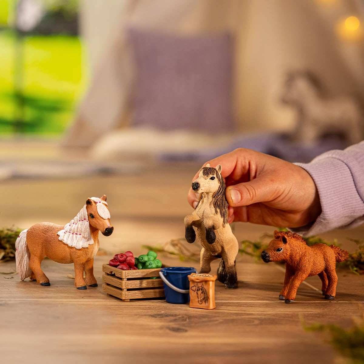 Hand eines Kindes, die mit kleinen Spielzeugpferden neben winzigen Kisten und Eimern auf einem Holzboden spielt.