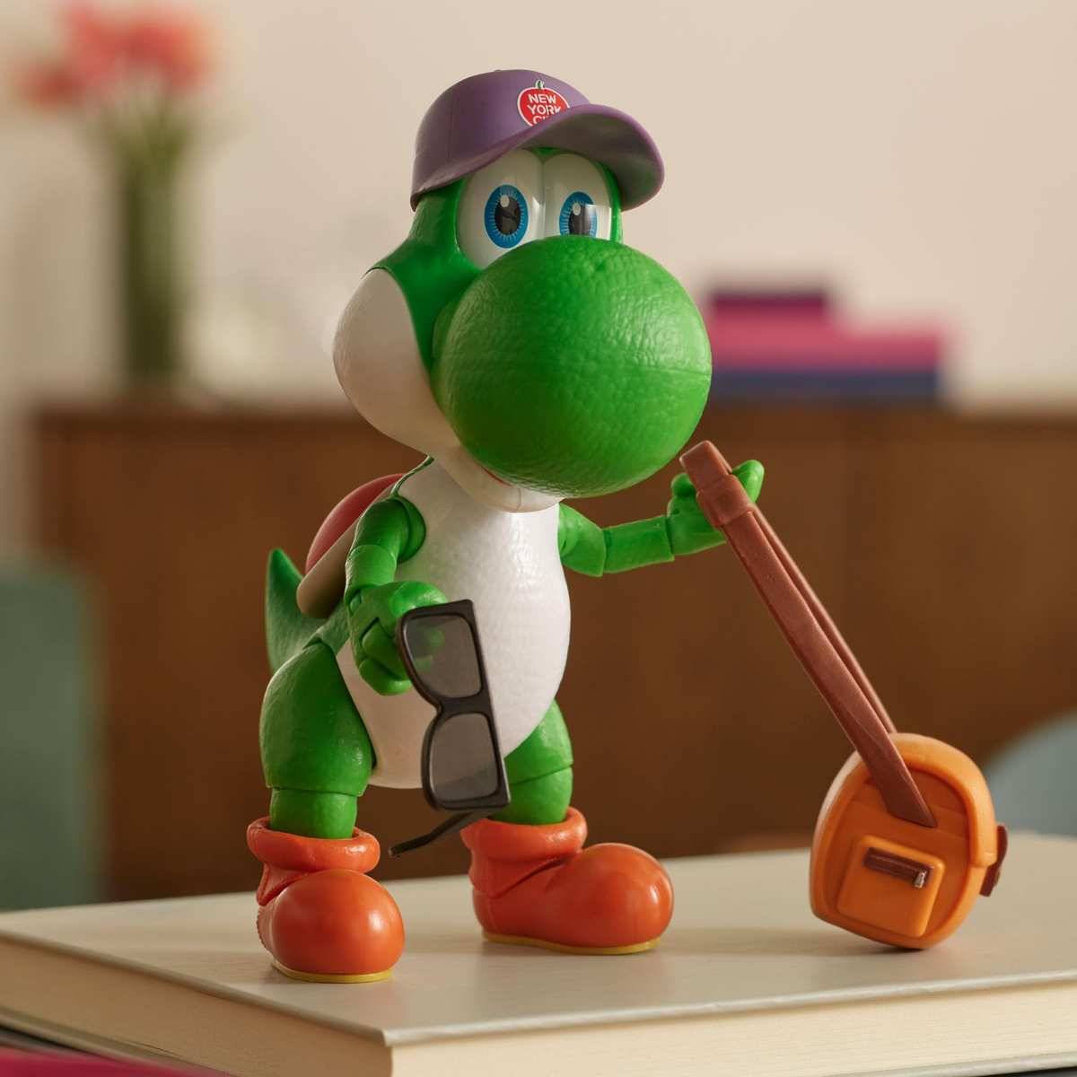 Yoshi-Spielzeugfigur mit Mütze und Sonnenbrille in der Hand, lehnt sich an einen orangefarbenen Rucksack auf einem Buch.