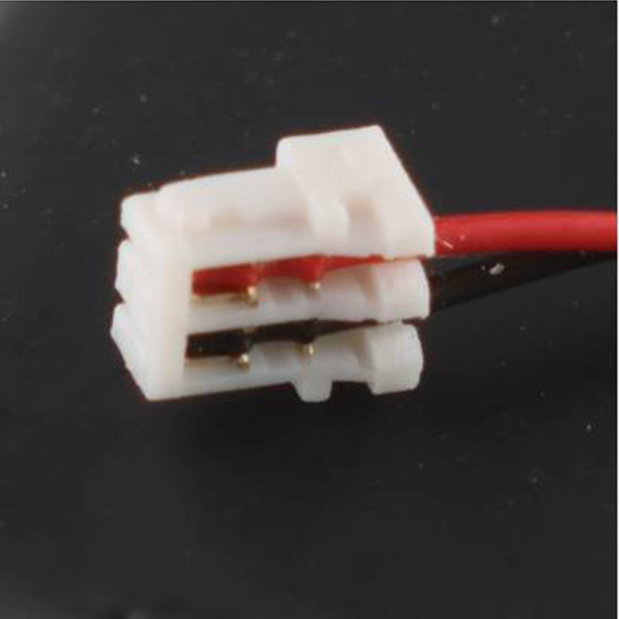 Nahaufnahme eines kleinen weißen zweipoligen Elektrosteckers mit angeschlossenen roten und schwarzen Drähten vor dunklem Hintergrund.