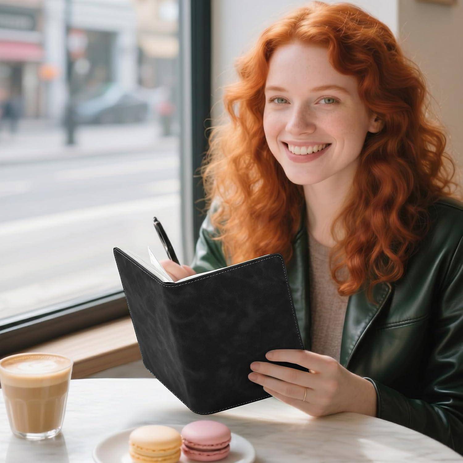 Eine lächelnde rothaarige Frau schreibt in einem Notizbuch an einem Cafétisch neben einem Latte und zwei Macarons neben einem Fenster.
