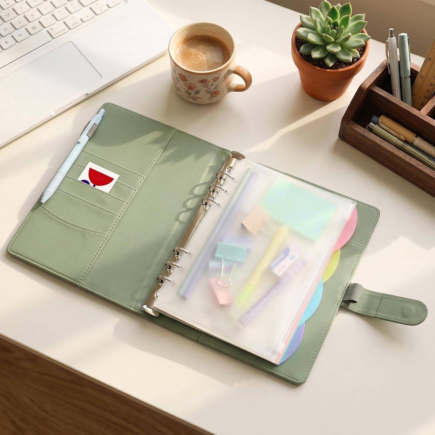 Offener grüner ringgehefteter Planer auf einem Schreibtisch, mit Pastellnotizen und Reitern in einer klaren Tasche neben einer Kaffeetasse, einer Sukkulente, einem Laptop und einem Stifthalter.