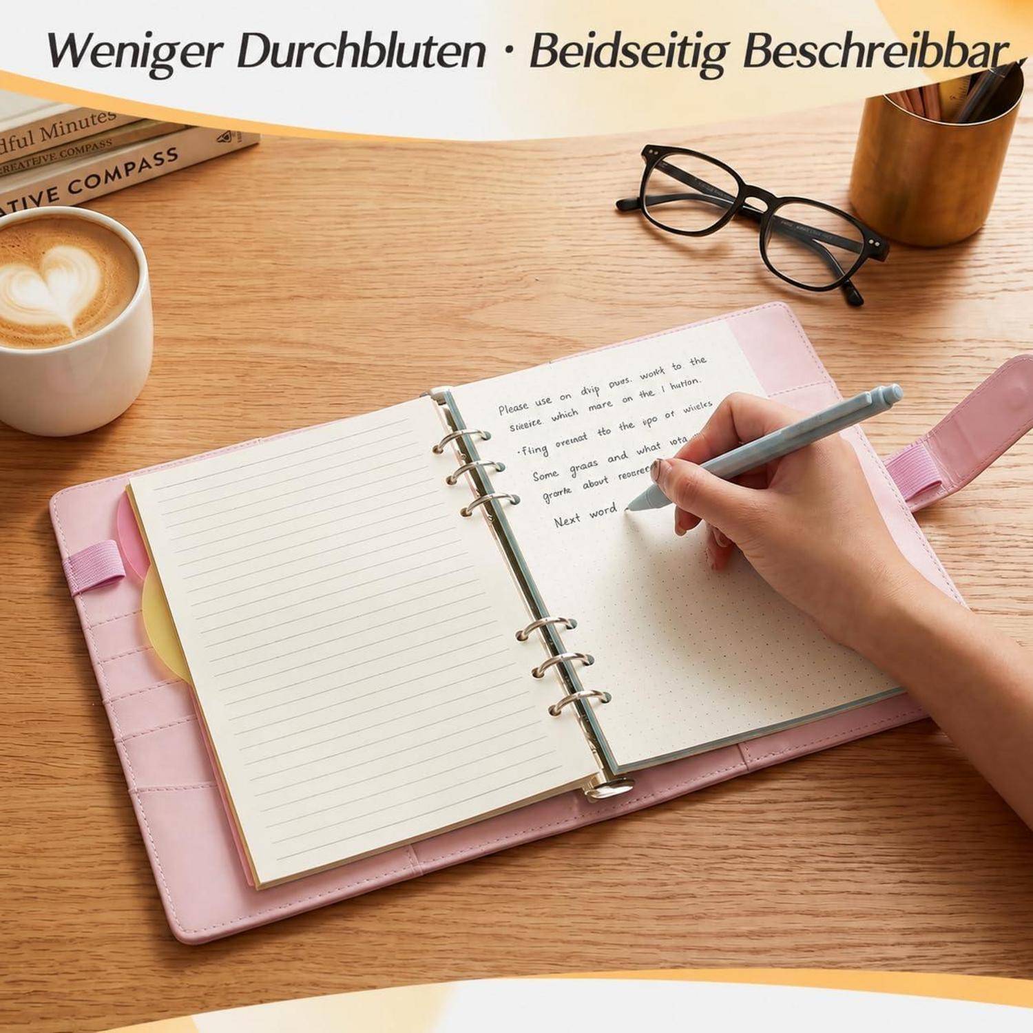 # Handschrift in einem rosa spiralgebundenen Notizbuch auf einem Holzschreibtisch mit Kaffee, Brille und einem Stiftbecher; der Text sagt „Weniger Durchbluten · Beidseitig Beschreibbar