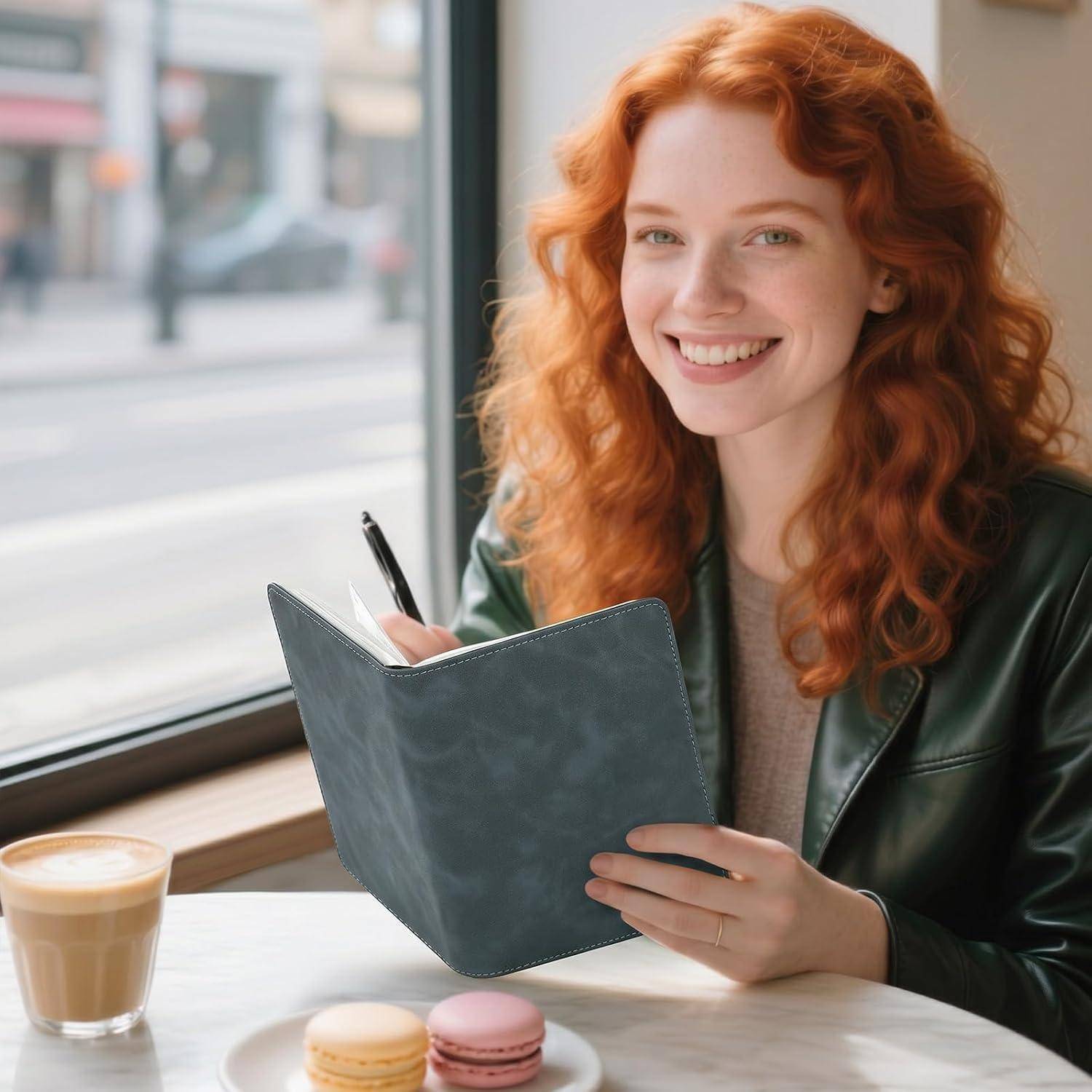 Lächelnde rotaarige Frau schreibt in einem Notizbuch an einem Cafétisch neben einem Latte und zwei Macarons am Fenster.