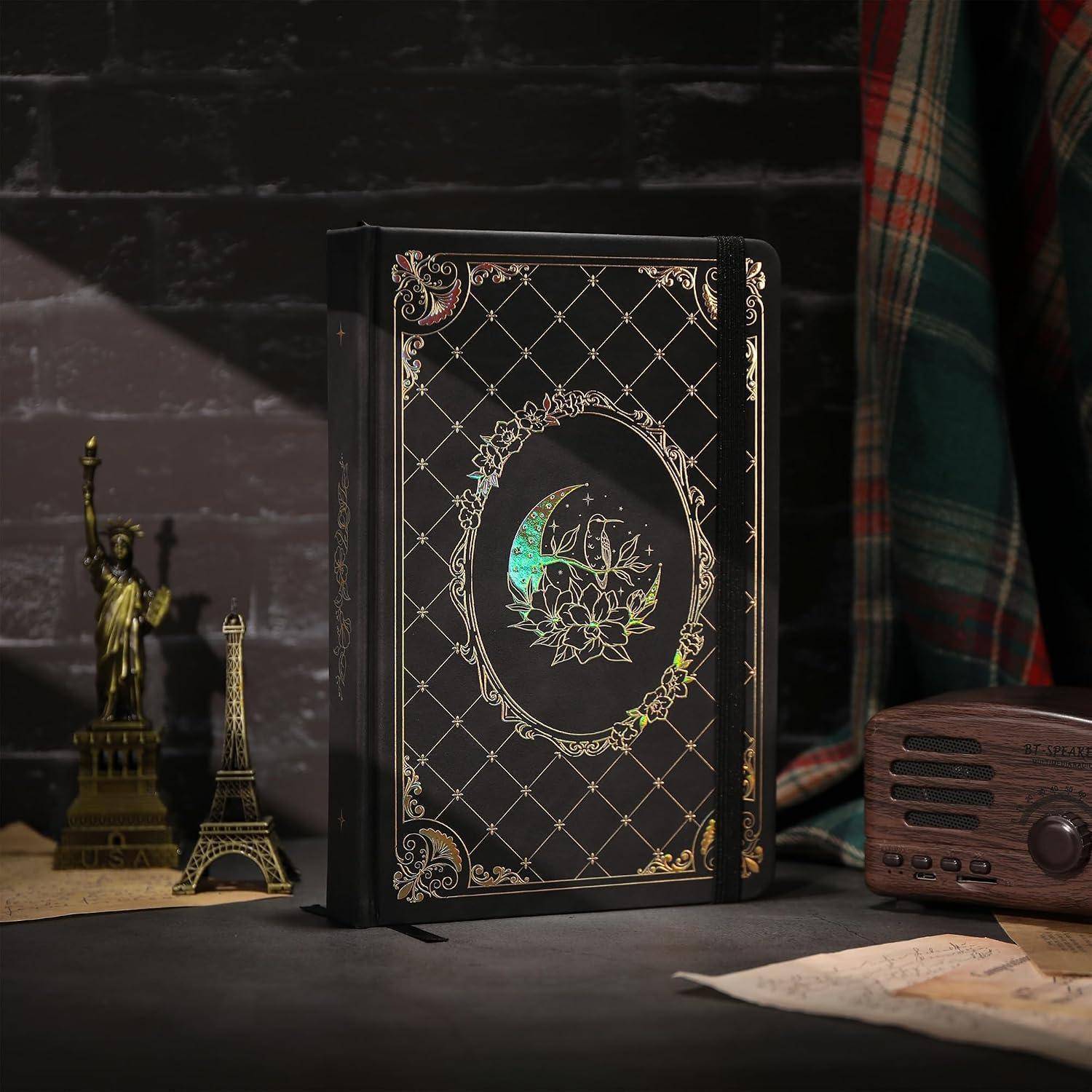Schwarzes verziertes Hardcover-Buch mit goldener Gitterwerk- und Blumenmondgestaltung, gezeigt mit Eiffelturm- und Freiheitsstatuen-Figuren auf einem Schreibtisch.