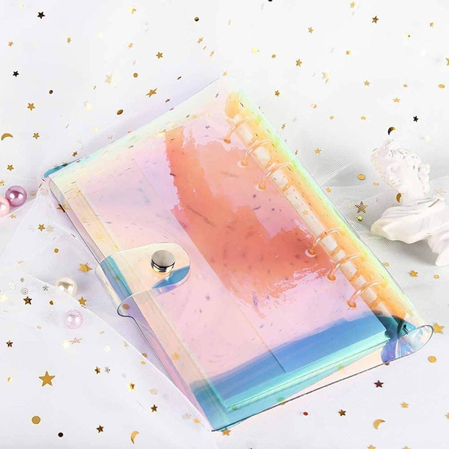 Pastellfarbenes Notizbuch mit irisierendem Spiralbindung und durchscheinendem Einband, das auf einer weißen Oberfläche liegt, die mit goldenem Sternchenconfetti verstreut ist.