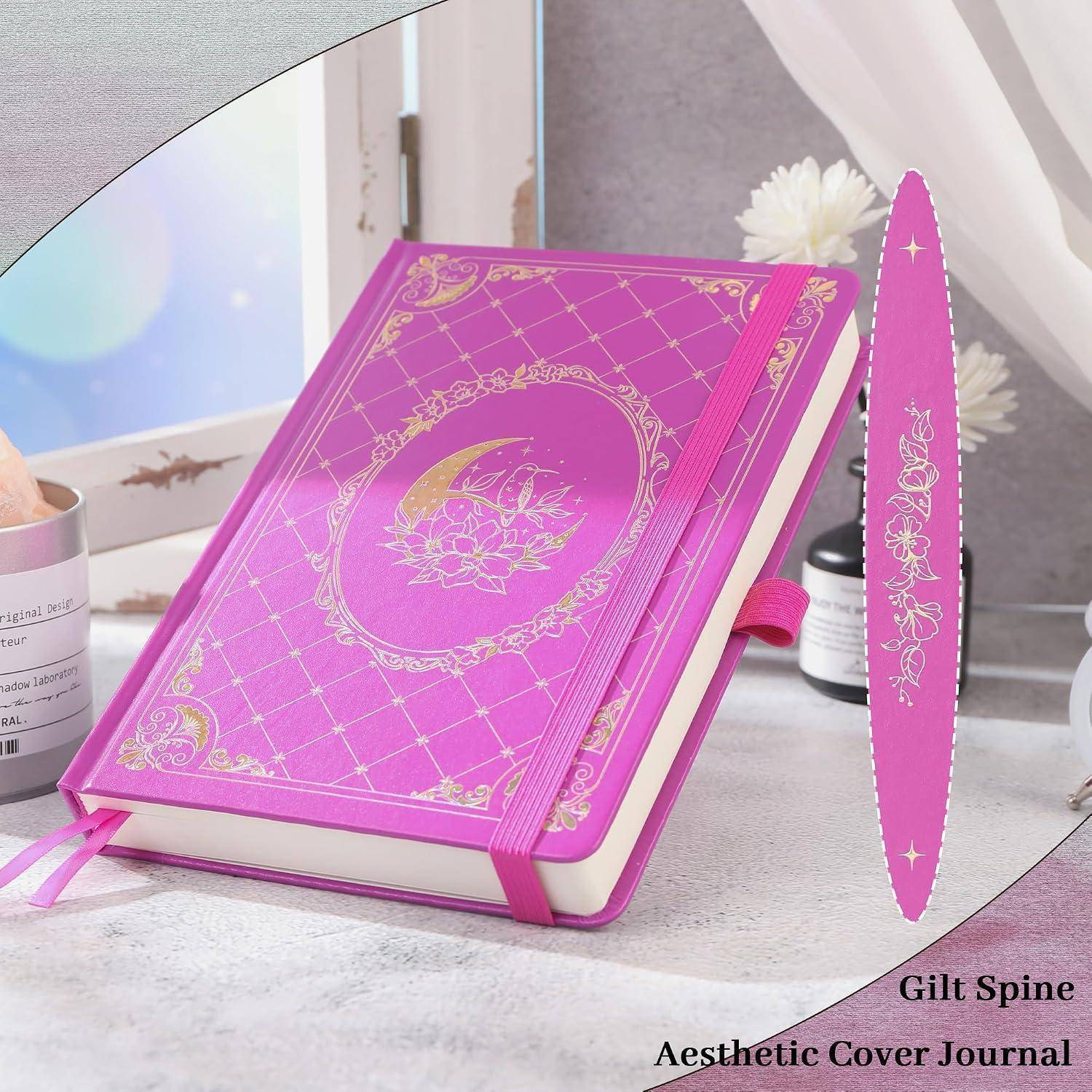 Rosa Hardcover-Tagebuch mit goldenem dekorativem Filigree, Gummiband und vergoldetem Rücken, das auf einem Schreibtisch ausgestellt ist.