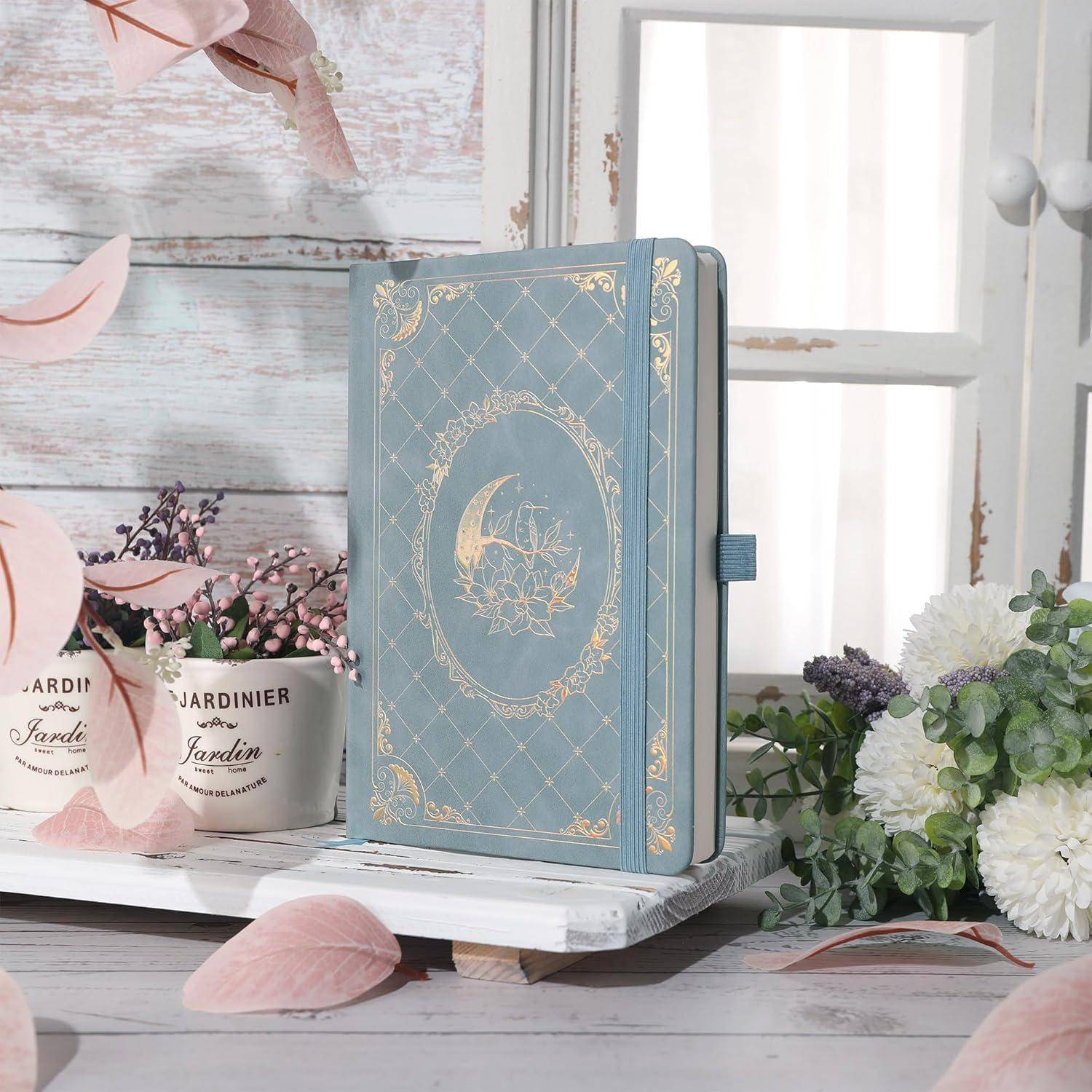 Dekoratives blaues Hardcover-Tagebuch mit goldenem Blumendesign auf einem weißen Regal, umgeben von rosa Blütenblättern und Topfblumen in einer hellen, rustikalen Umgebung.